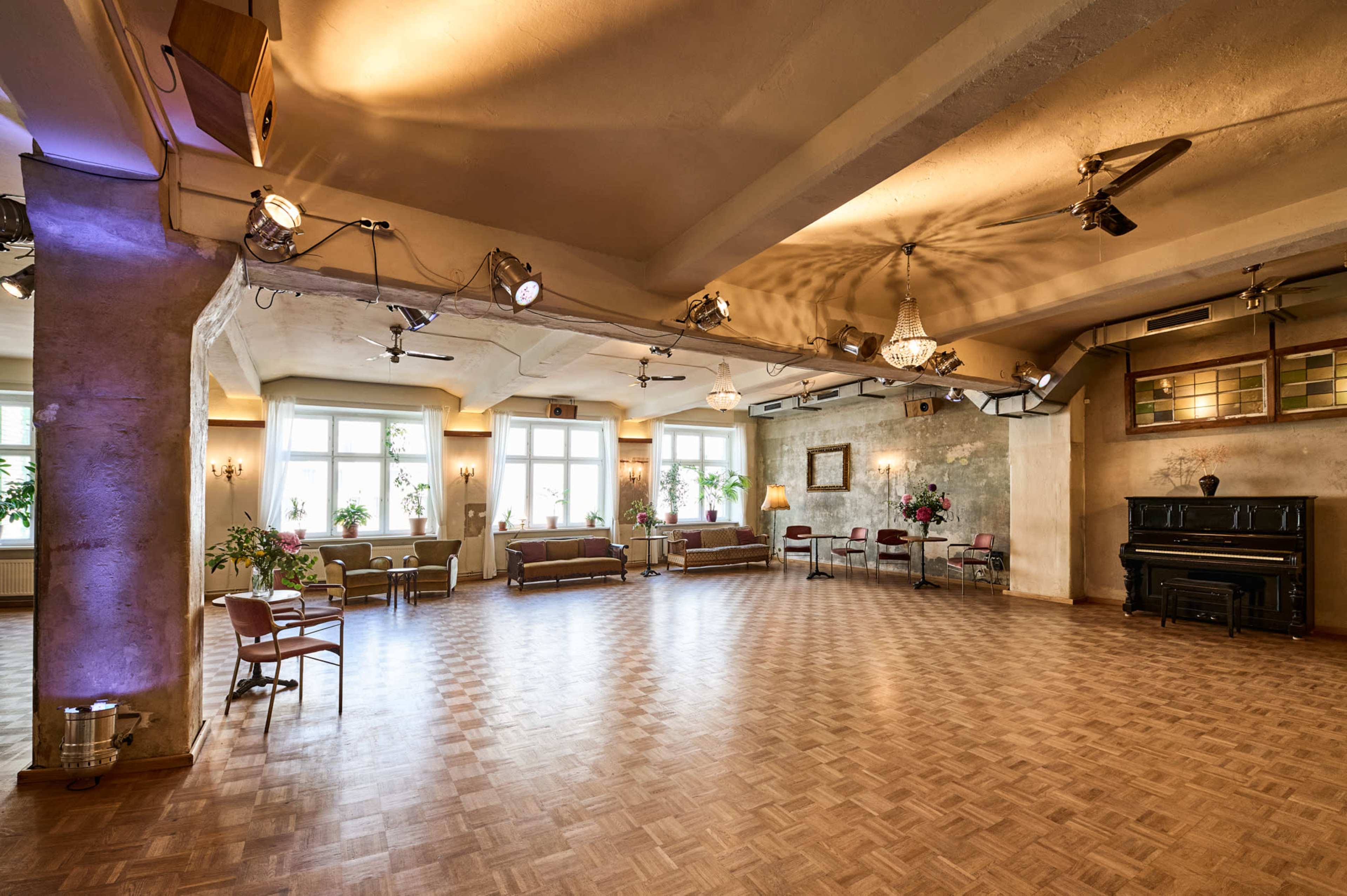 A spacious indoor event space with wooden flooring, large windows, and plants, featuring seating areas and a fireplace.
