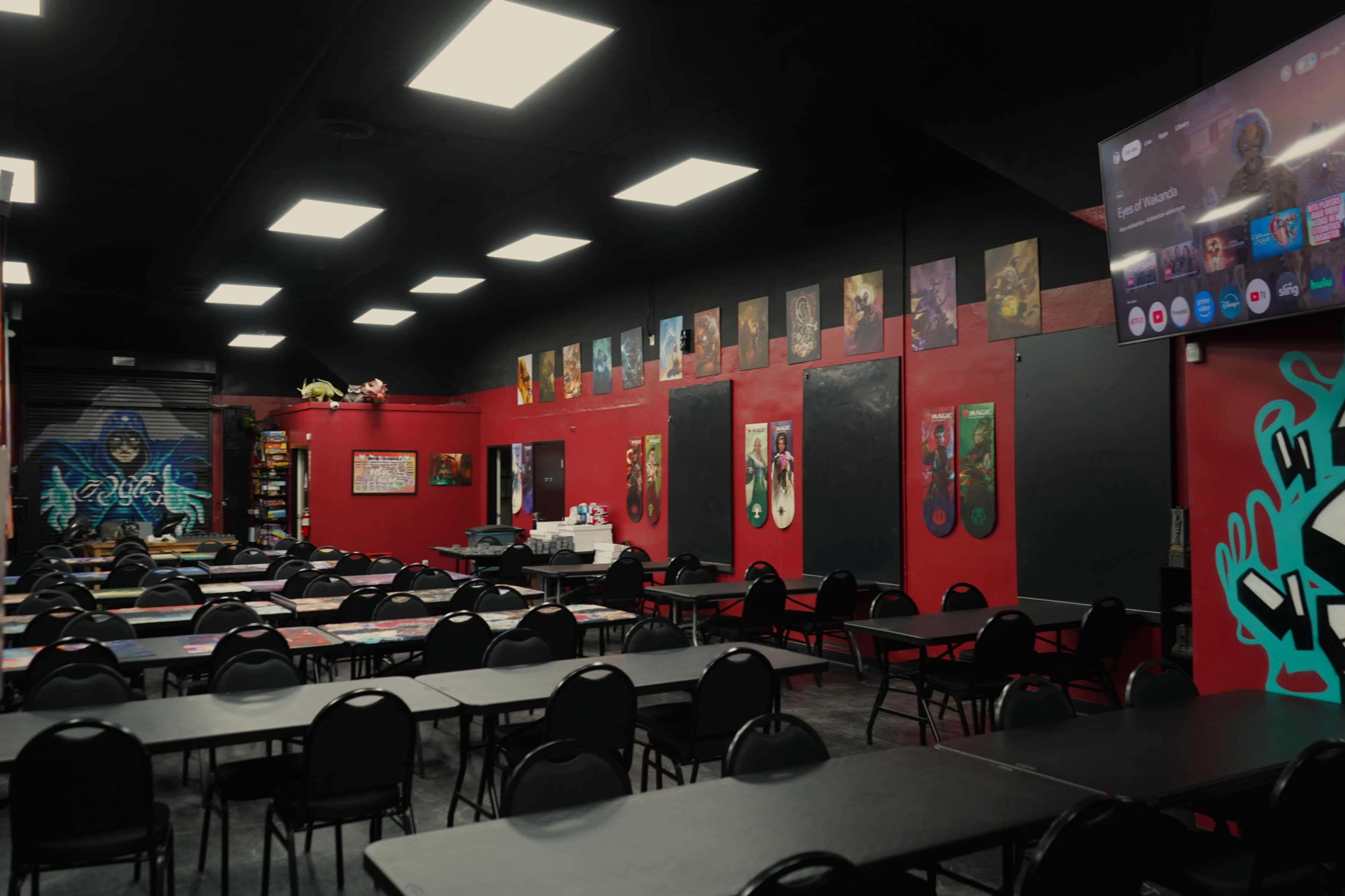 The image shows an empty room with multiple tables and chairs arranged for an event, featuring colorful wall art and a large screen on one wall.