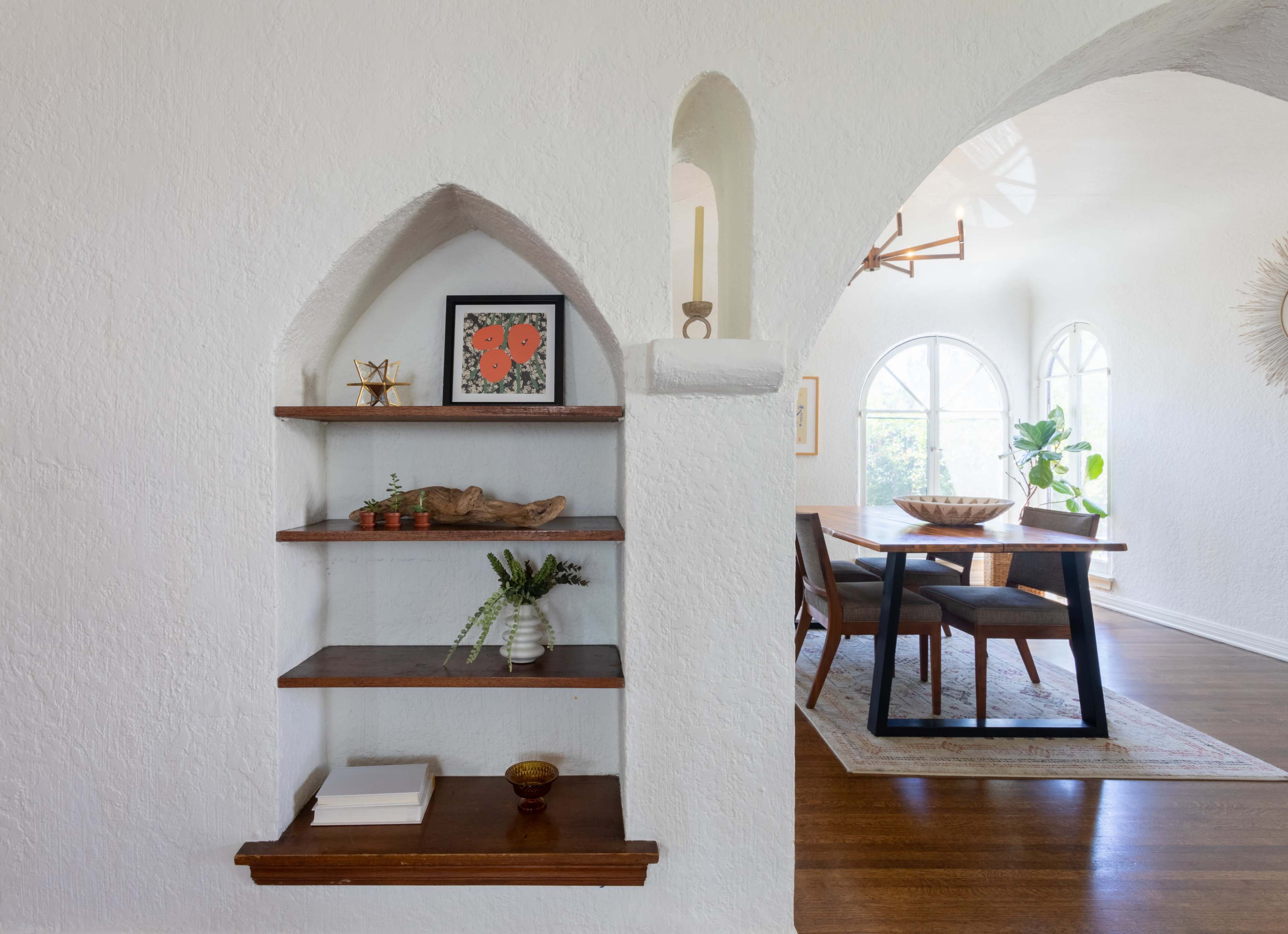A white-walled alcove with wooden shelves displaying decorative items, leading to a dining area with a wooden table and chairs.