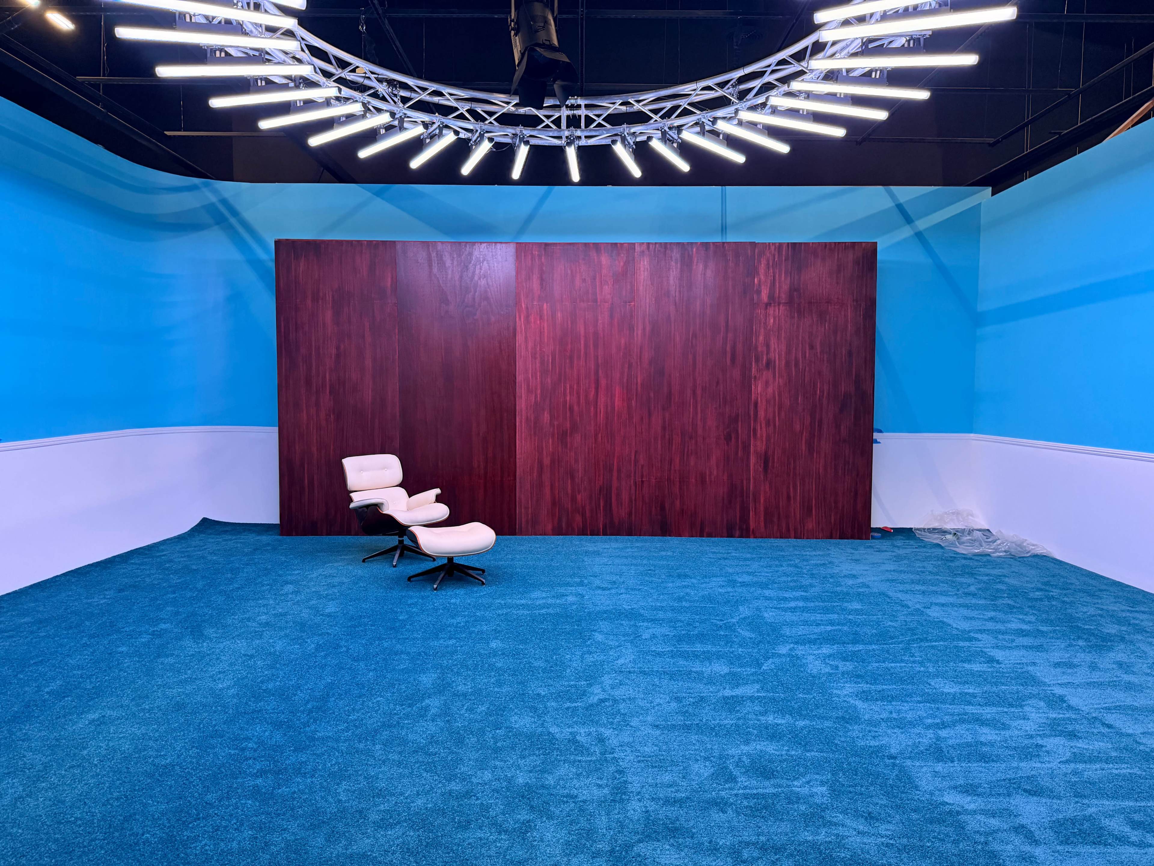 The image shows an empty studio space with a teal carpet, a large wooden wall, and a white lounge chair positioned in front.