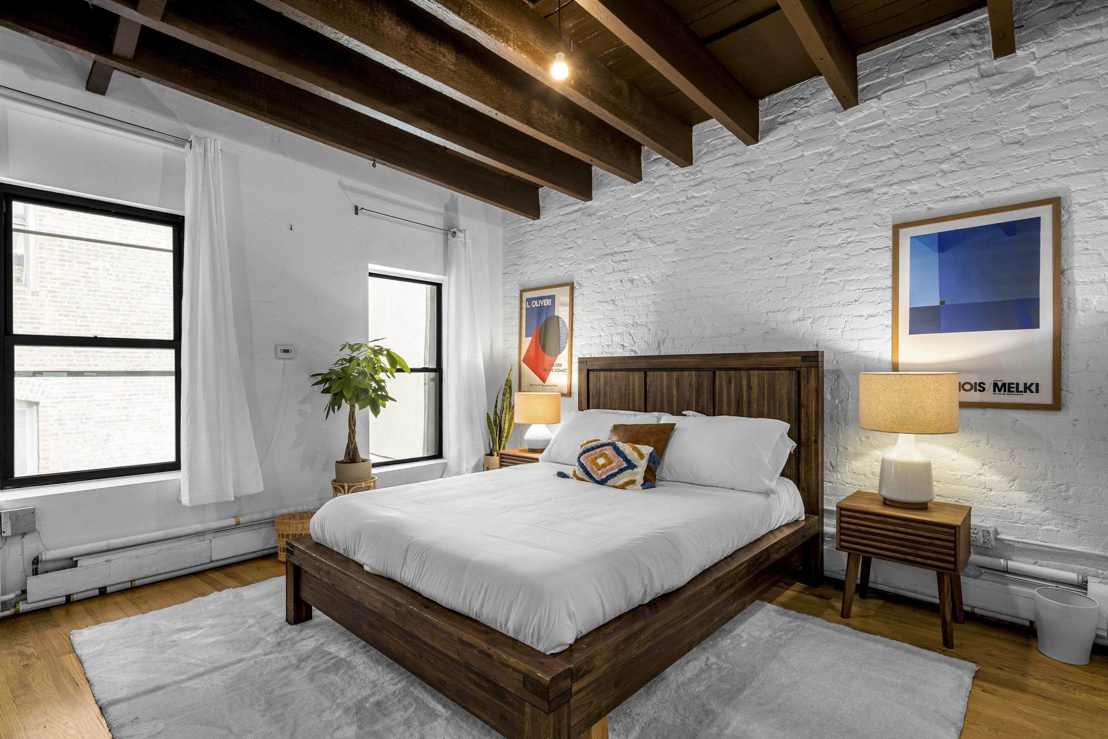 A modern bedroom with a wooden bed frame, white bedding, a potted plant, and artwork on the brick wall, illuminated by natural light from large windows.