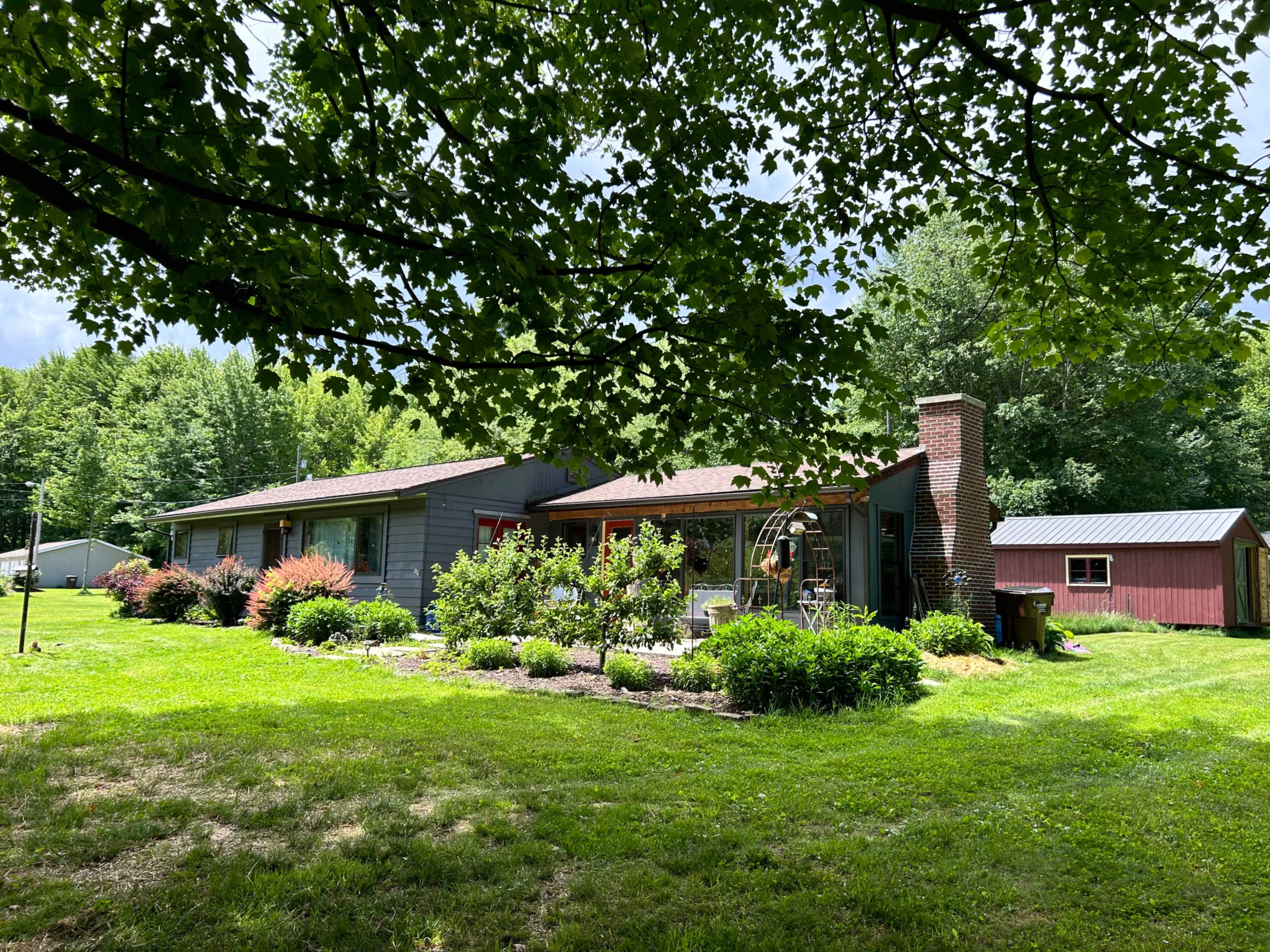 A single-story gray house with a red door and a brick chimney is surrounded by lush greenery and a small red shed in the background.