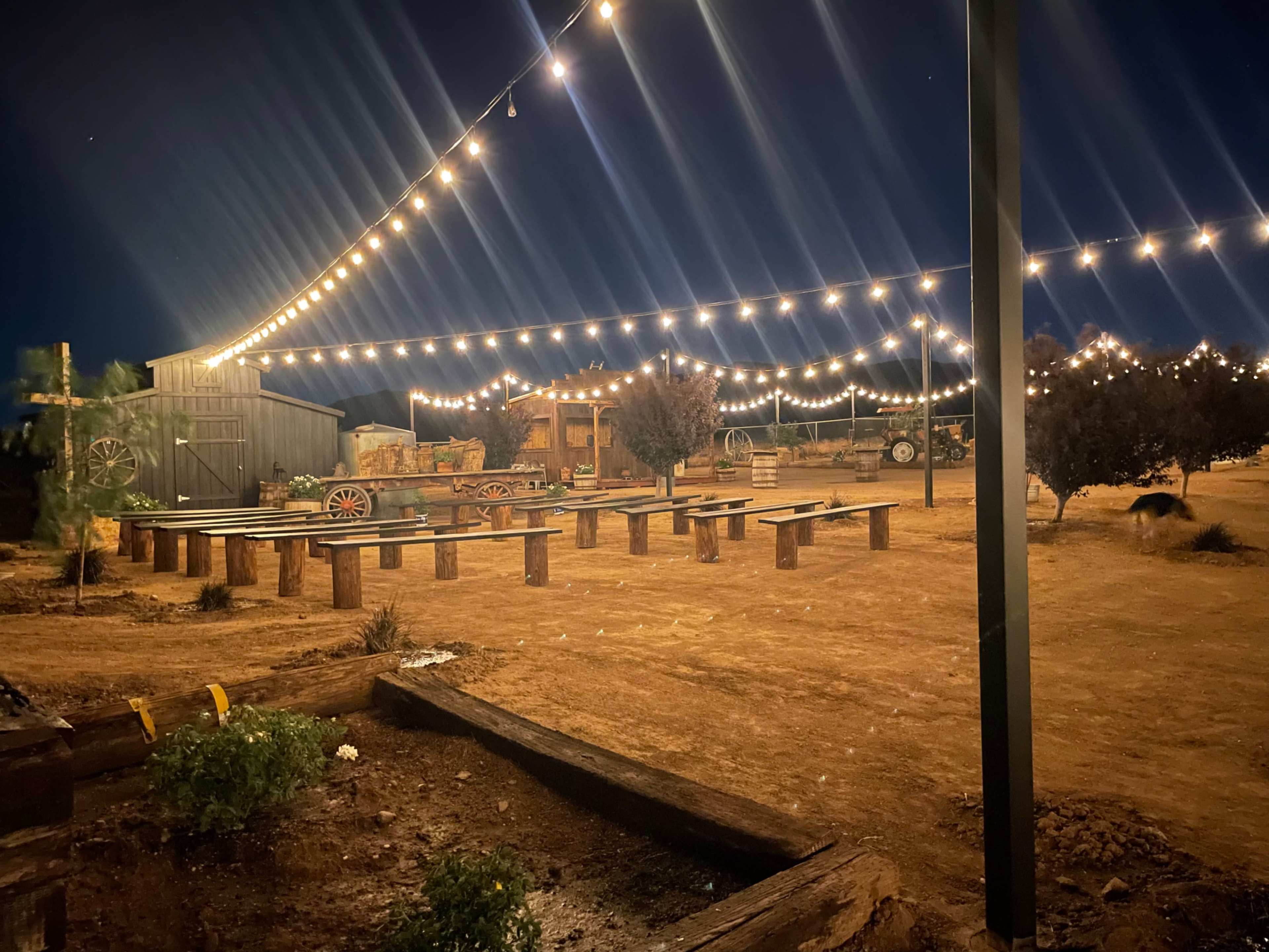 An outdoor event space lit by string lights, with wooden benches arranged in a dirt area surrounded by trees and rustic structures.