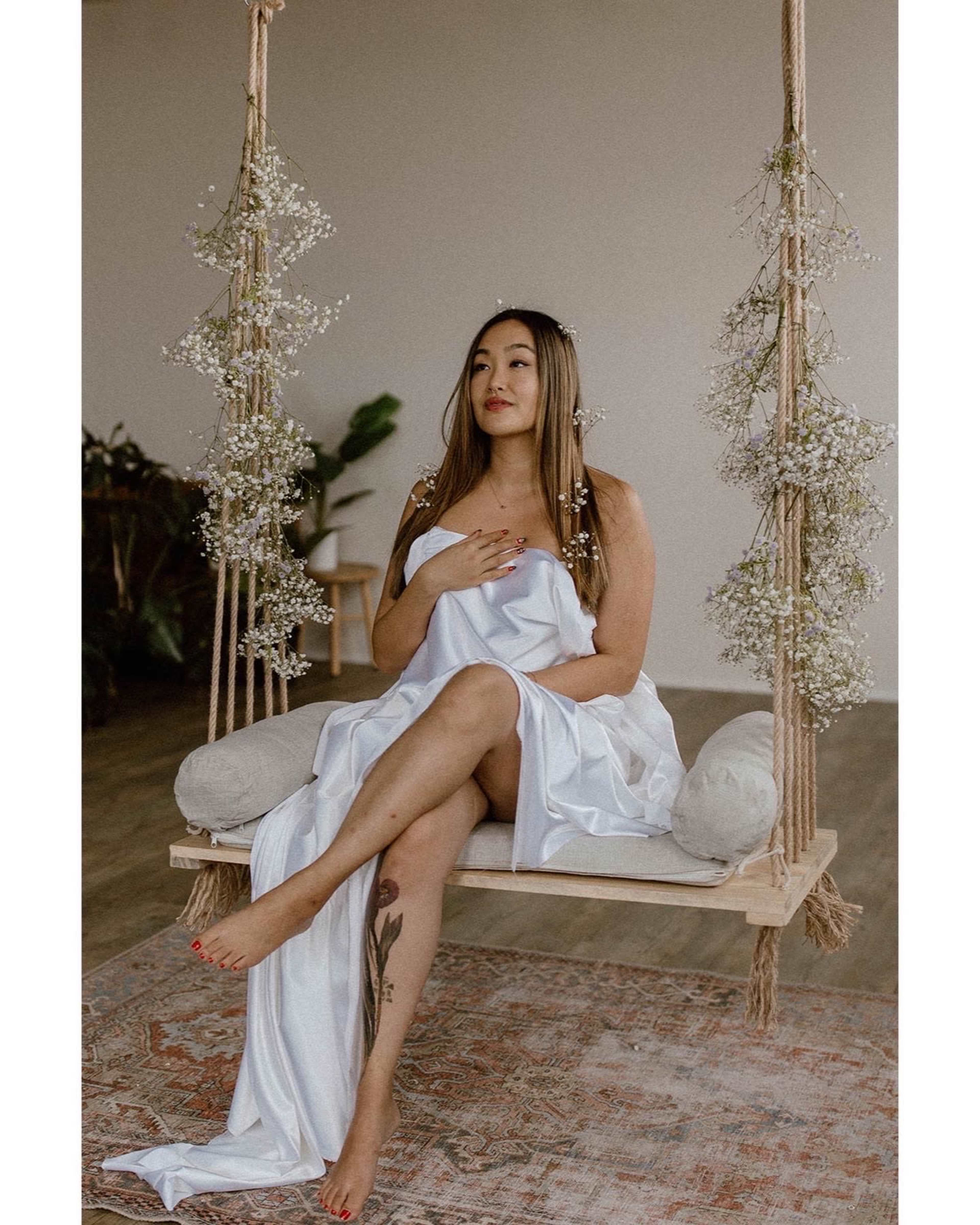 A woman sits on a wooden swing adorned with flowers, wearing a white dress, in a softly lit room.
