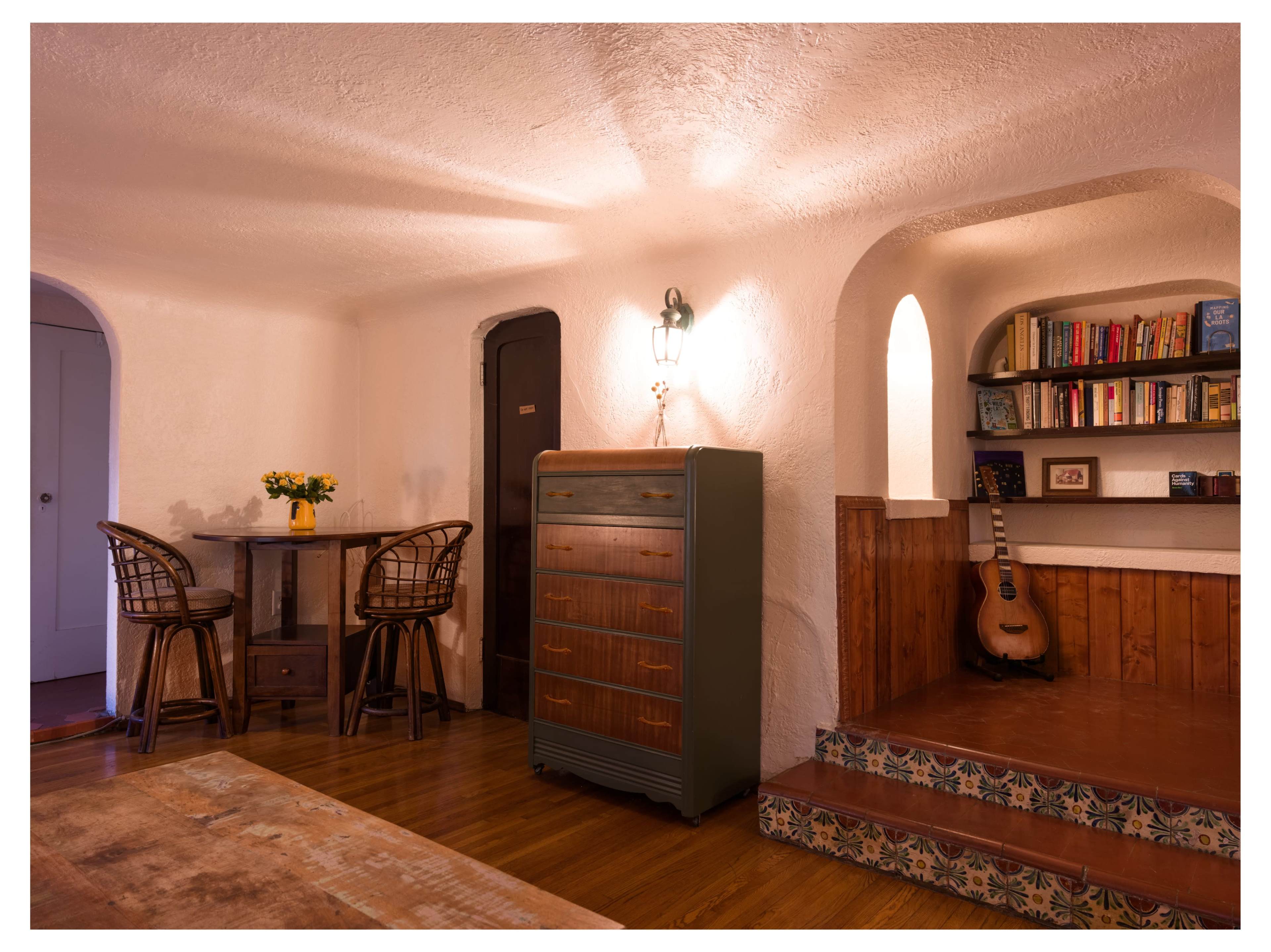 The image shows a cozy interior space with a small dining table, a chest of drawers, a bookshelf, and an acoustic guitar placed on a staircase.