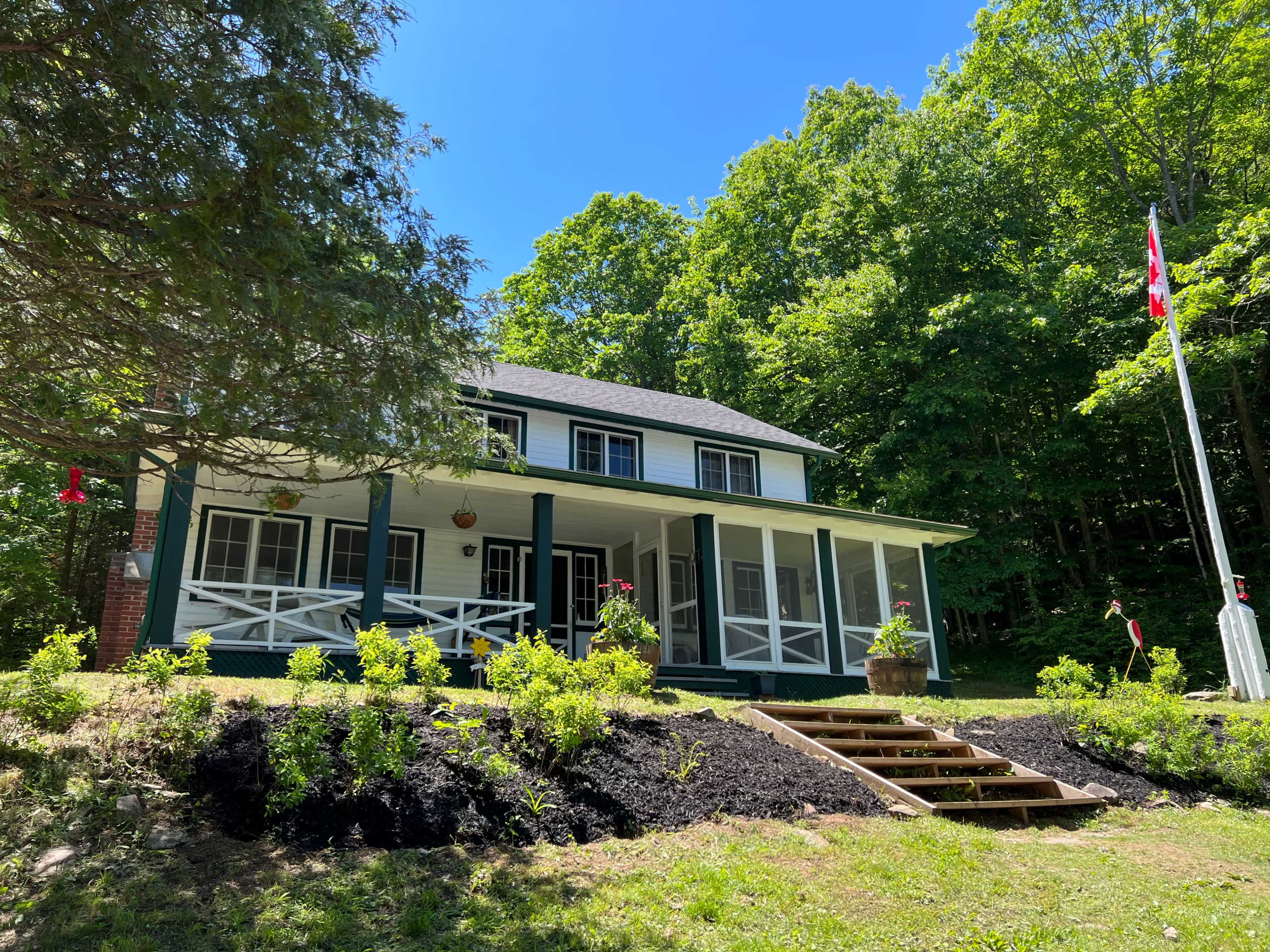 A two-story house with a porch is surrounded by trees and greenery, featuring steps leading up to the entrance and a flagpole.
