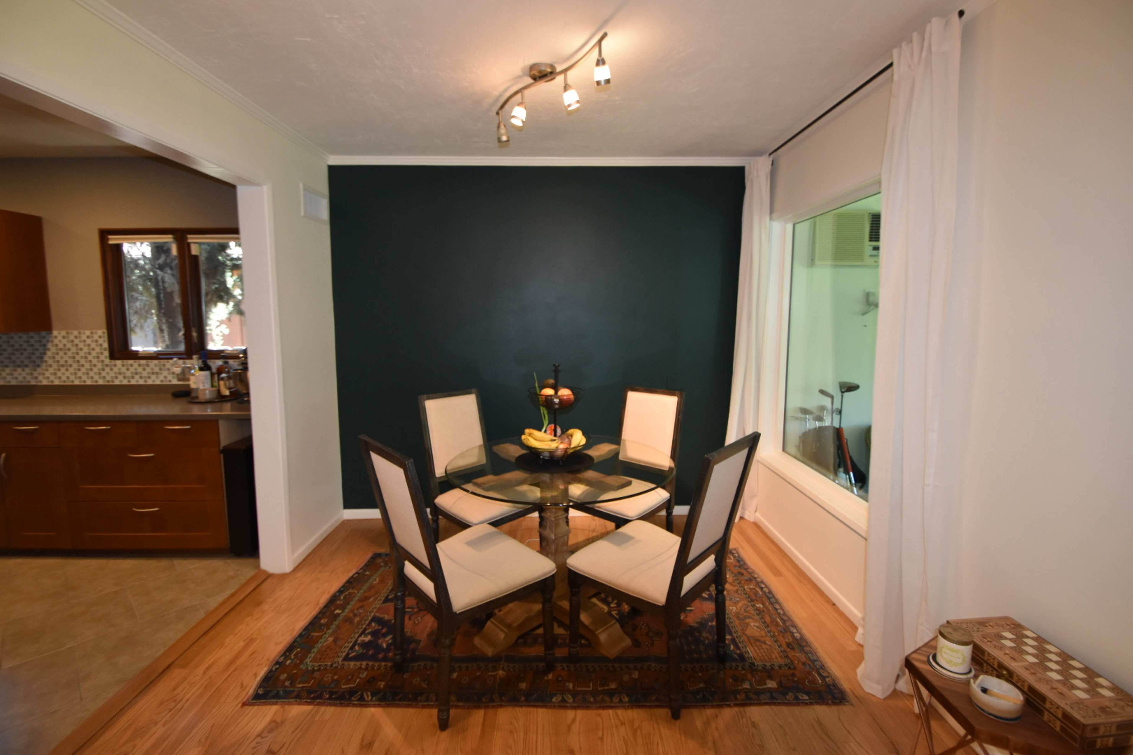 A glass dining table with four chairs is positioned against a dark green wall, with a window allowing light to enter from the adjacent room.