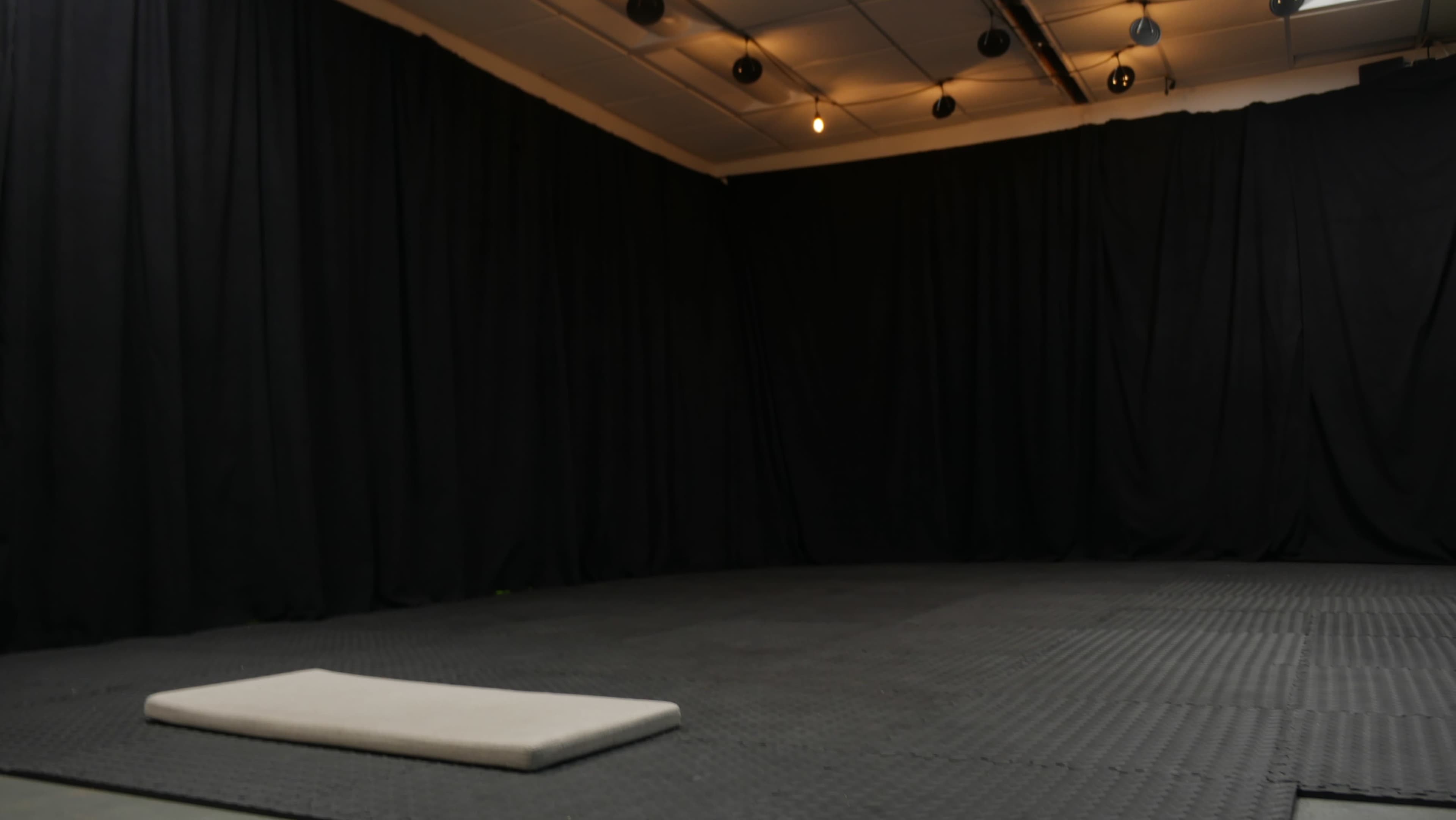 The image shows an empty room with black curtains, a padded mat on the floor, and overhead lighting.