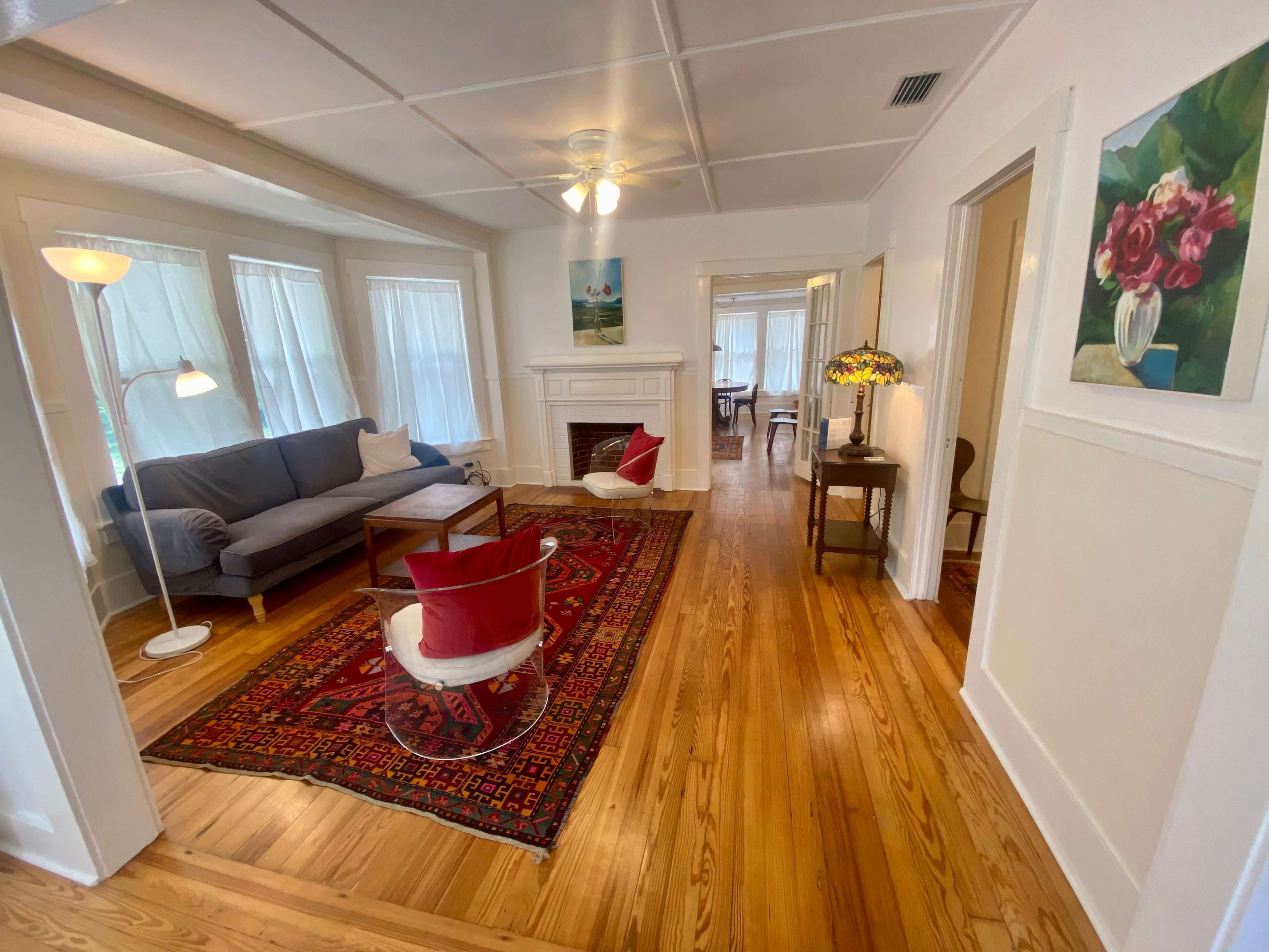 A bright, airy living room with a blue sofa, two red chairs, a lamp, and a large area rug, leading to adjacent spaces through open doorways.