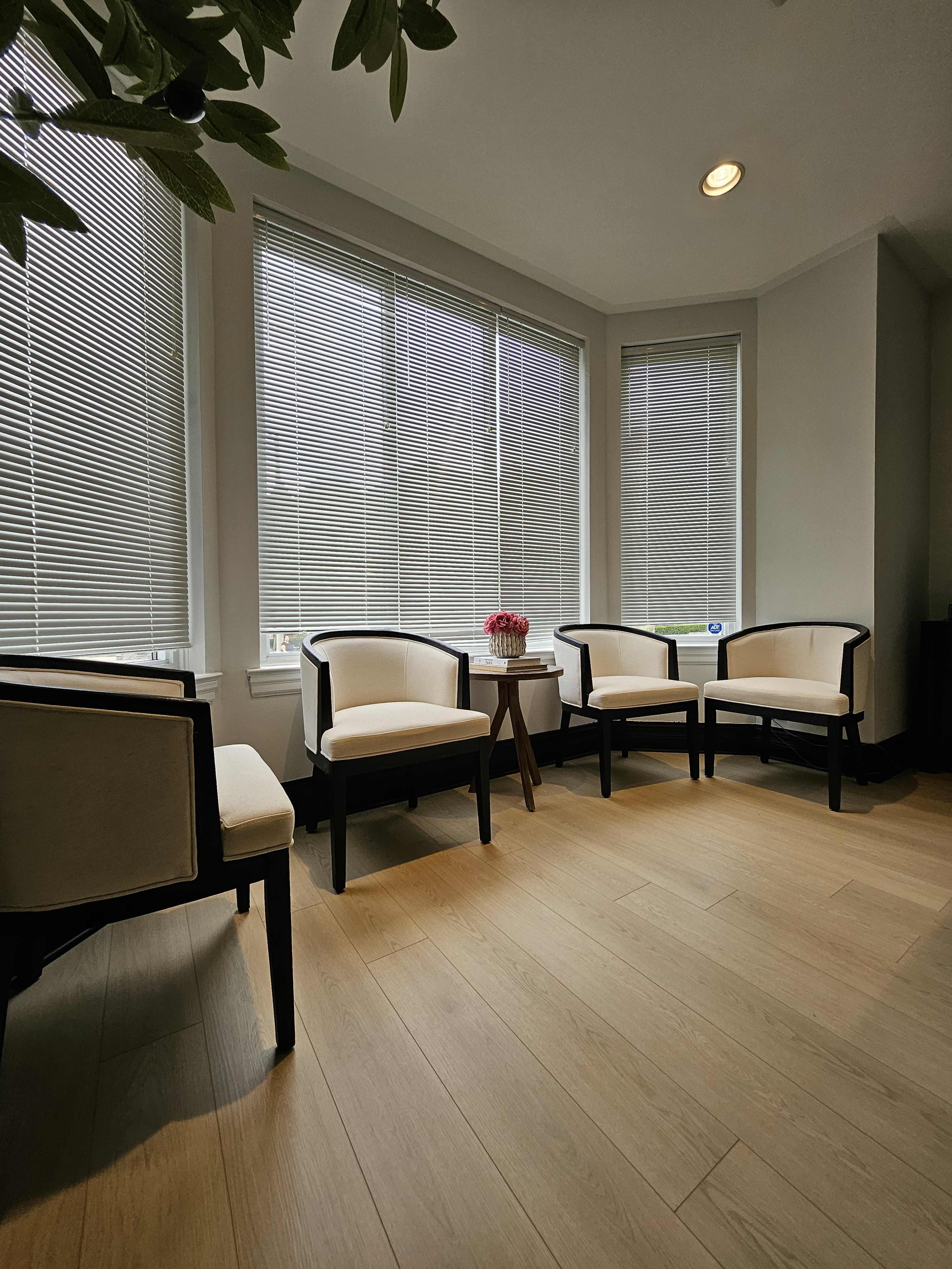 A seating area features four upholstered chairs arranged around a small table, illuminated by natural light filtering through window blinds.