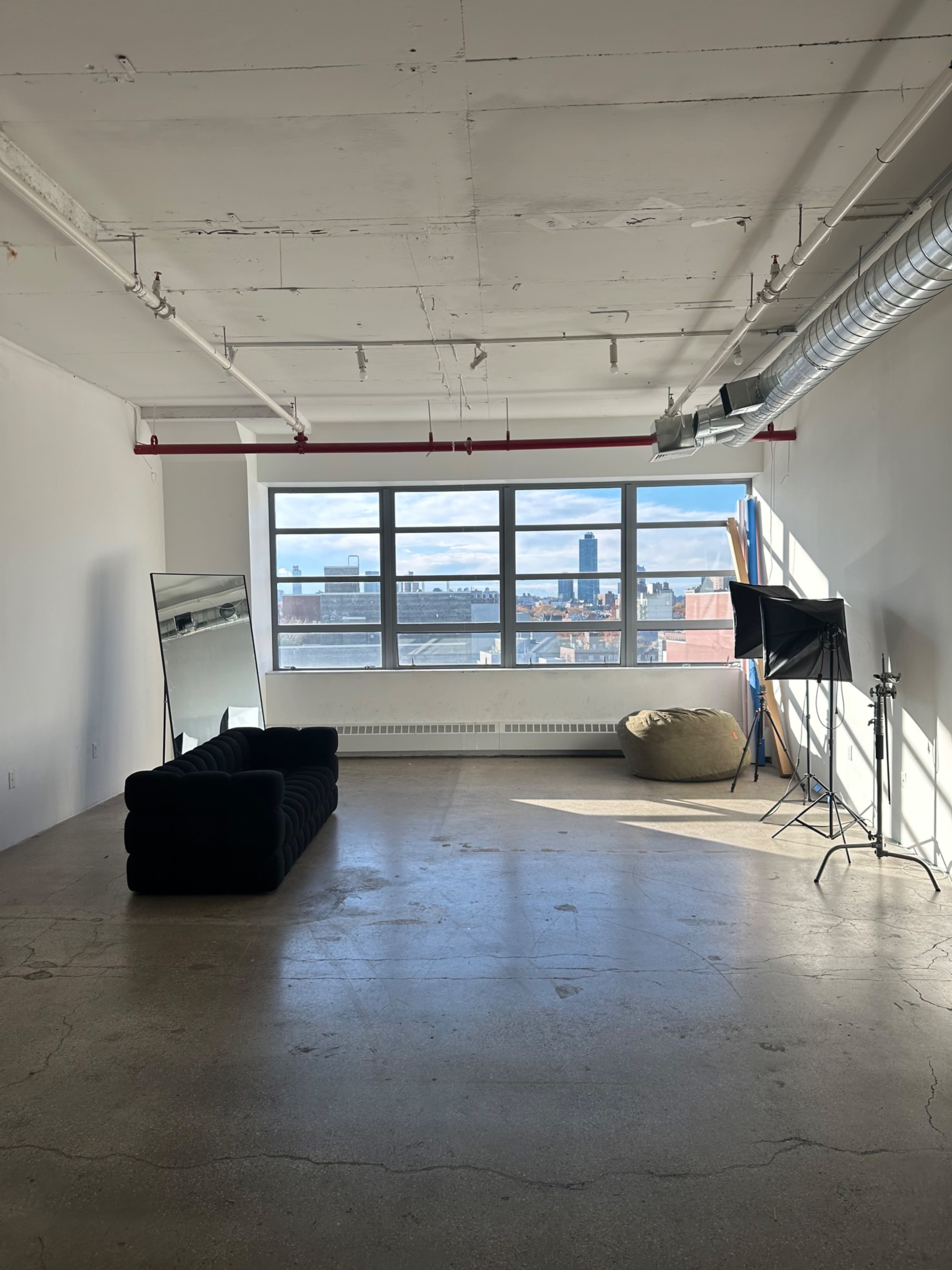 The image shows an empty room with a black couch, a large mirror, a soft seating beanbag, and a light setup, all facing large windows with a view of the skyline.