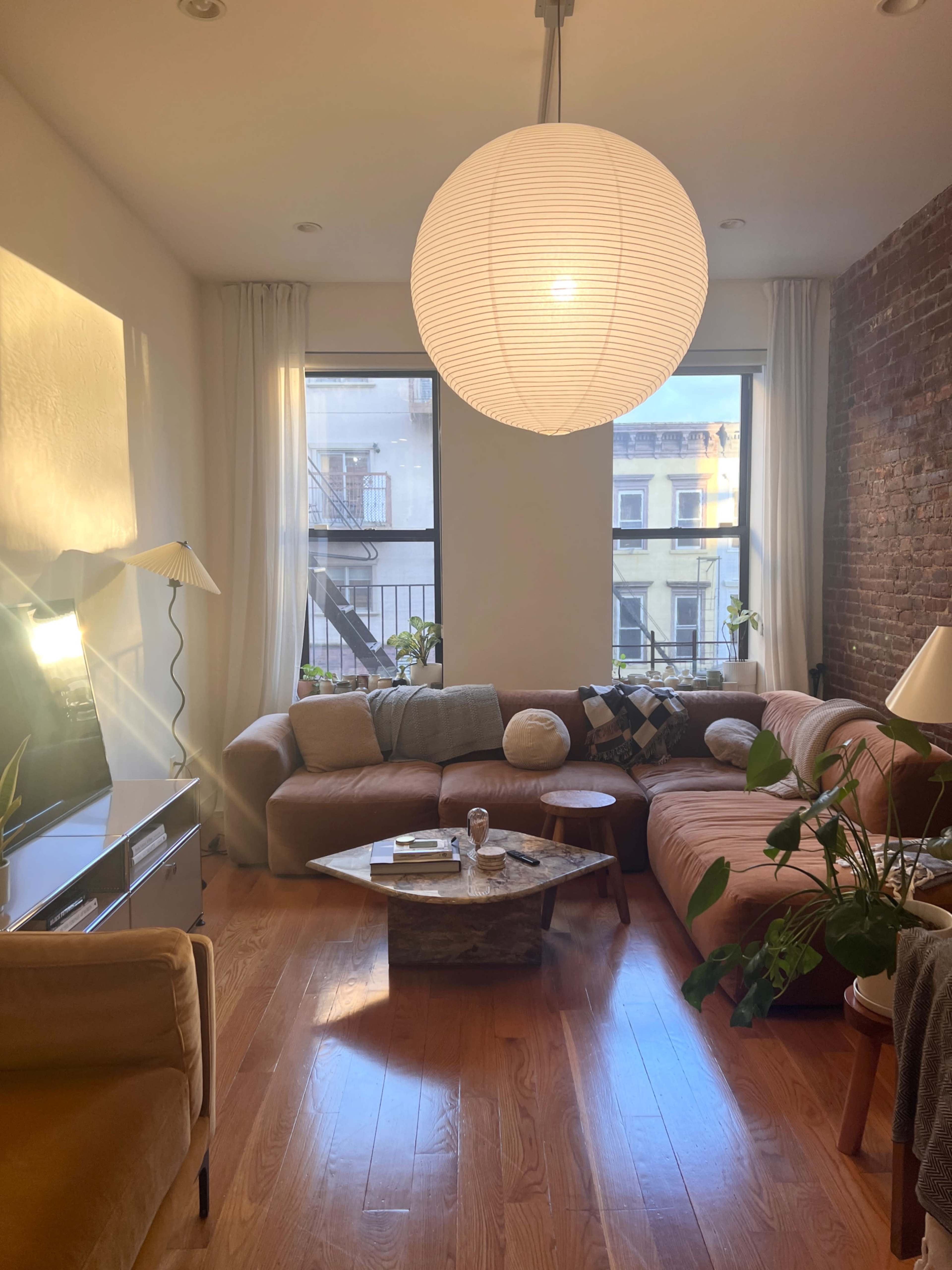 A living room features a sectional sofa, a coffee table, plants, and large windows with light streaming in.