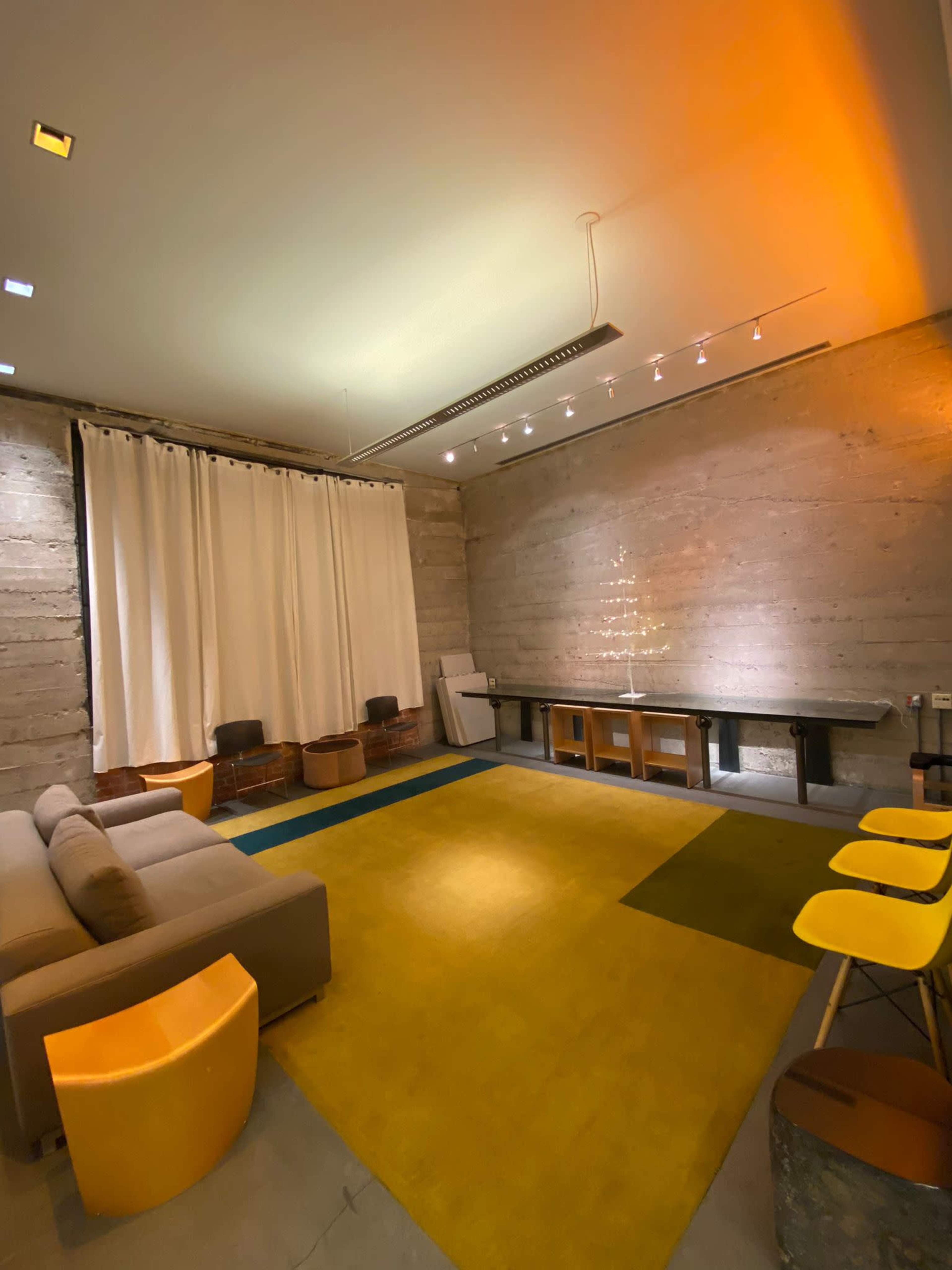 The room features a seating area with a sofa, yellow chairs, and a large rug, complemented by a concrete wall and soft curtains.