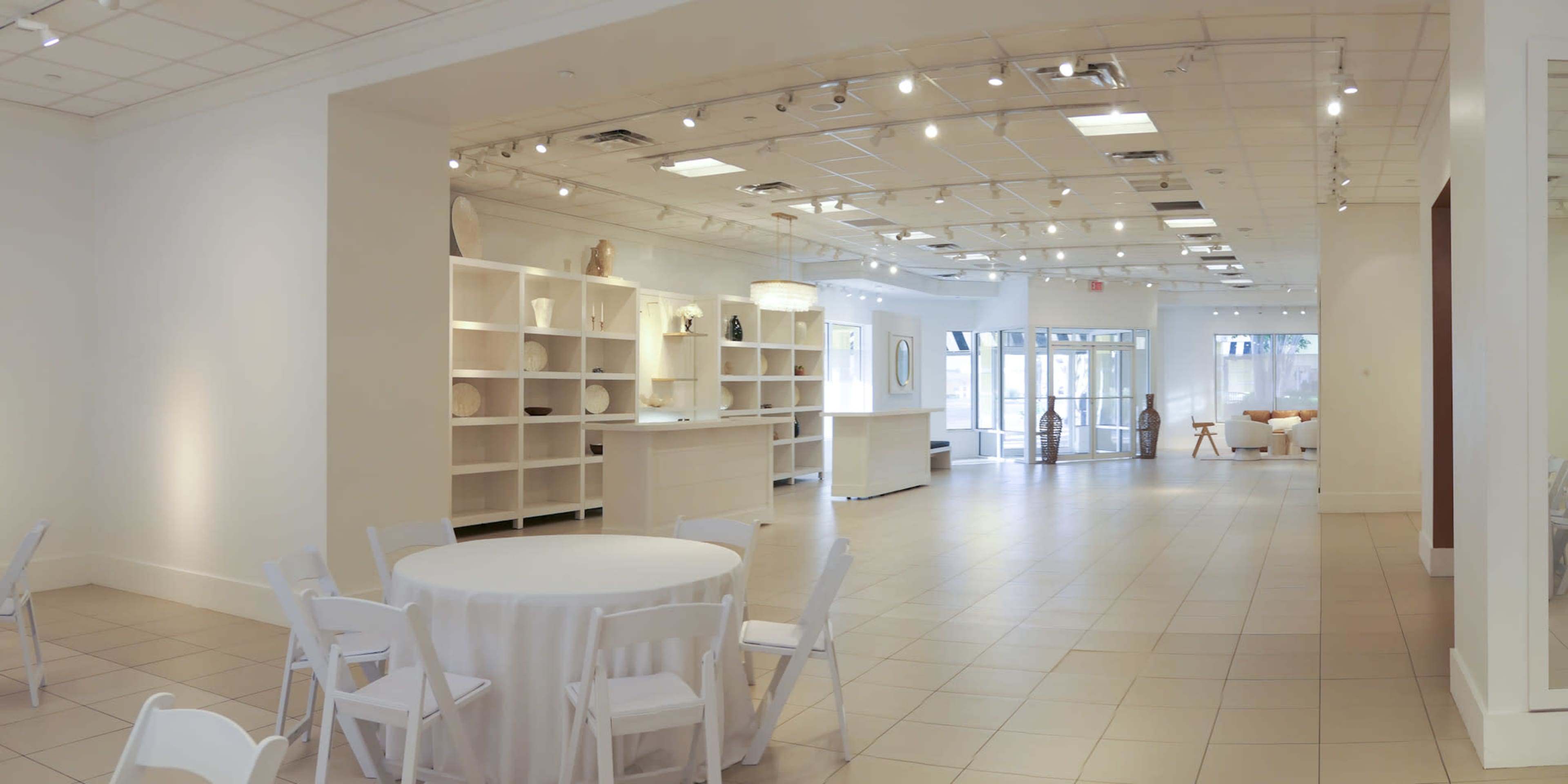The image shows a spacious, well-lit gallery or event space with white walls, a minimalist design, and a seating area featuring a circular table and chairs.