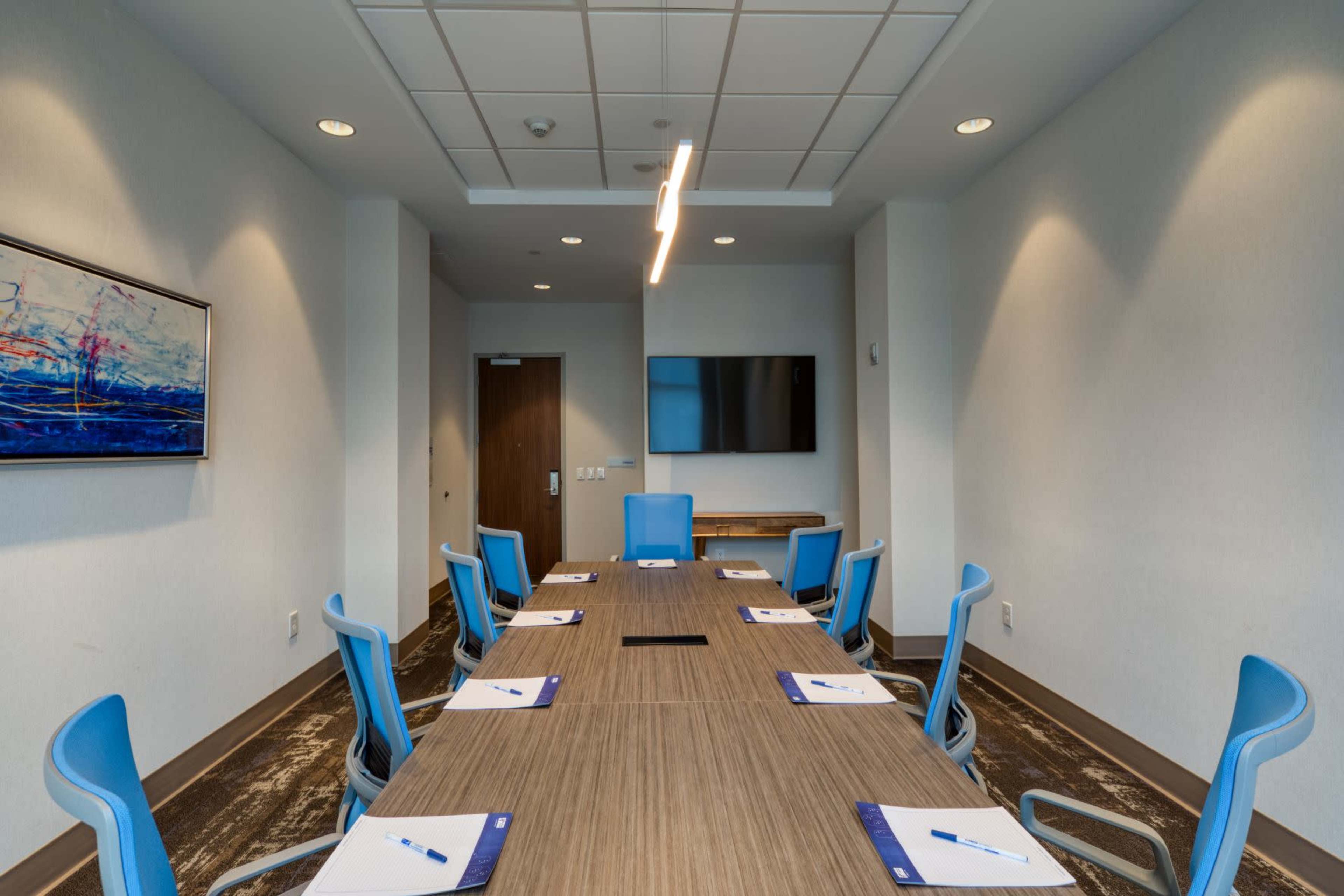 A modern conference room features a long wooden table surrounded by blue chairs, with a wall-mounted television and a piece of abstract artwork.