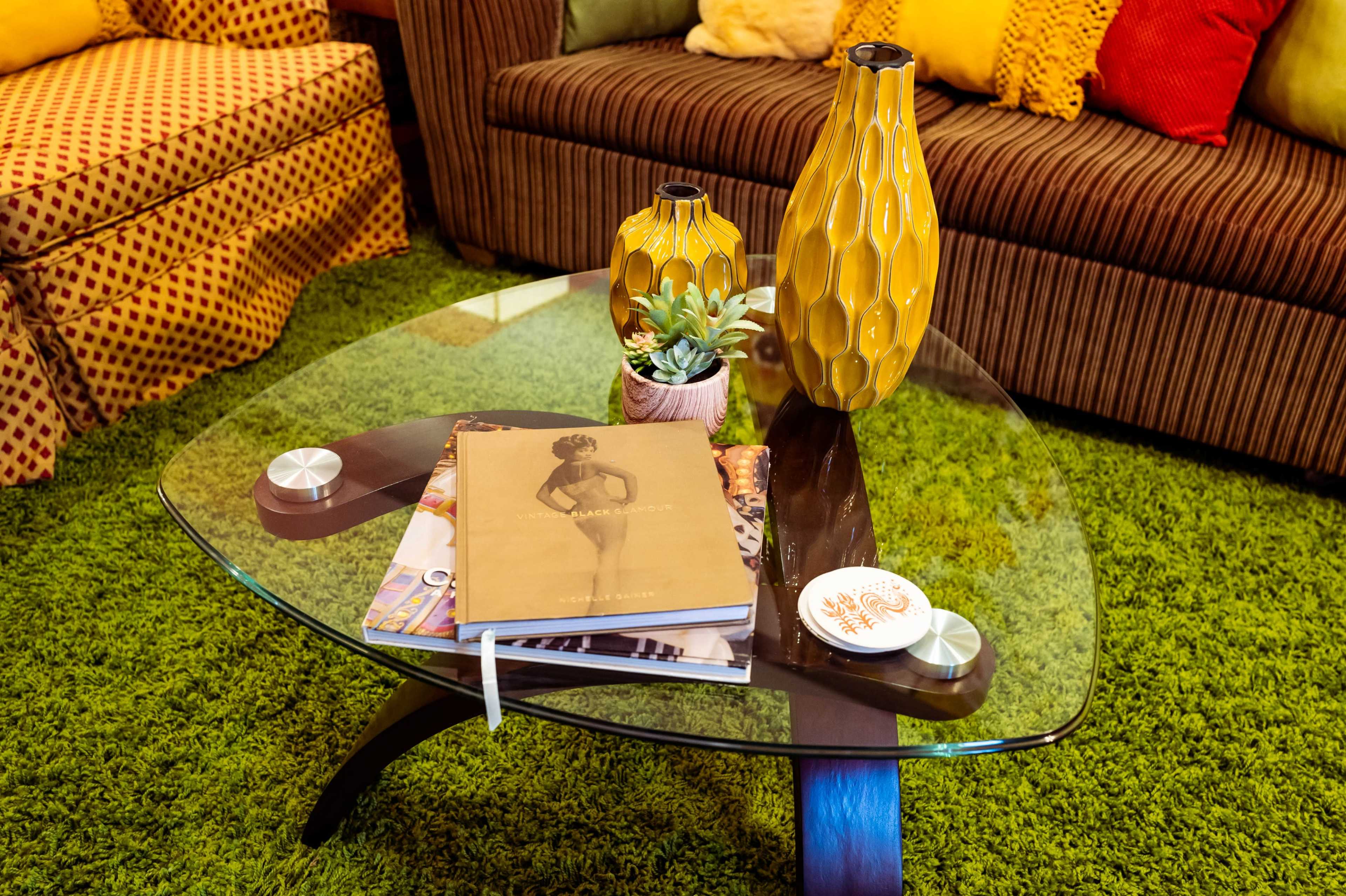 A glass coffee table with a wooden base displays a book, a small potted plant, and two yellow vases in a living room with green carpet and a patterned sofa.