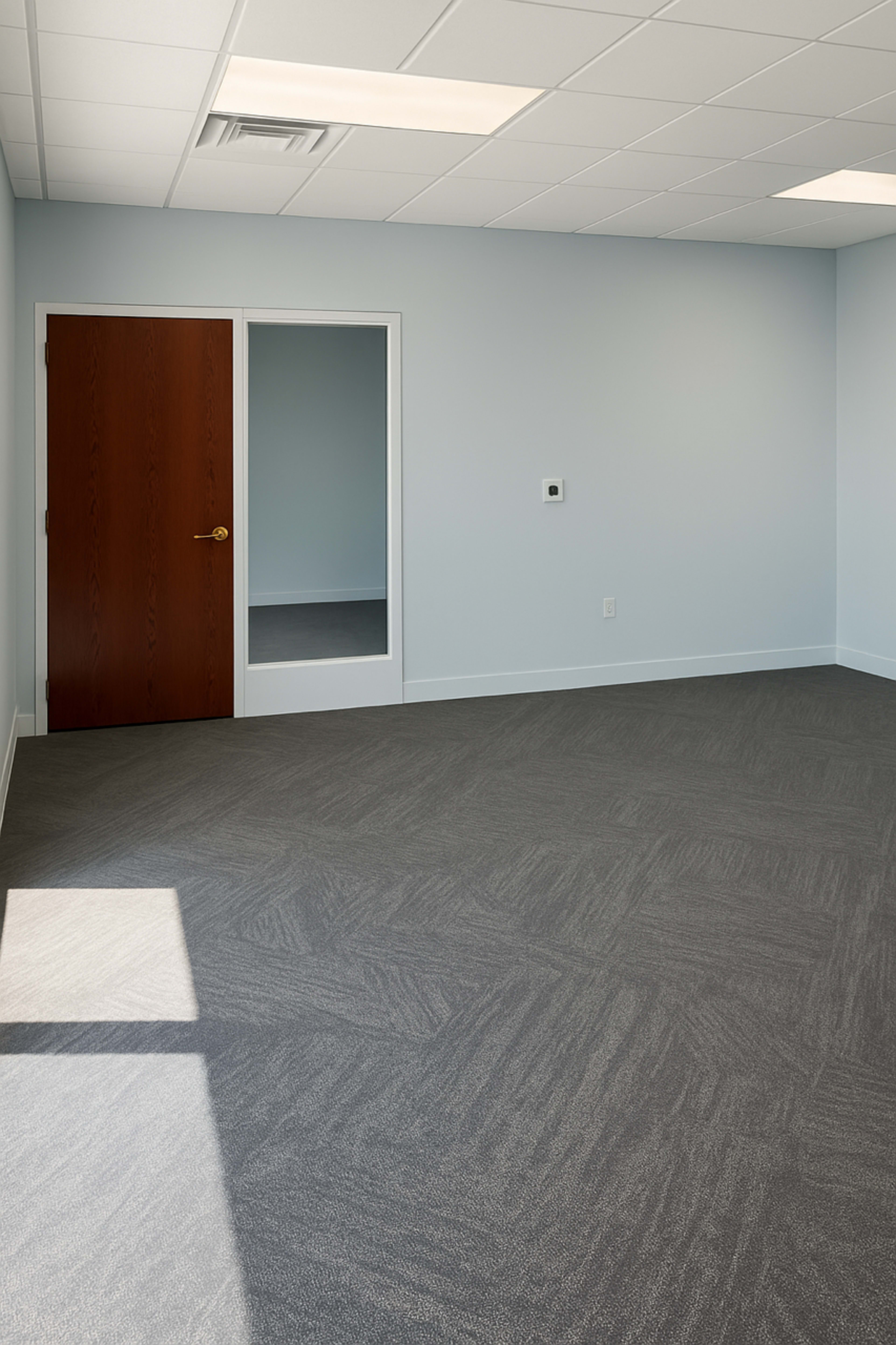 The image shows an empty office space with a large window, a brown wooden door, and a small mirror on the wall.