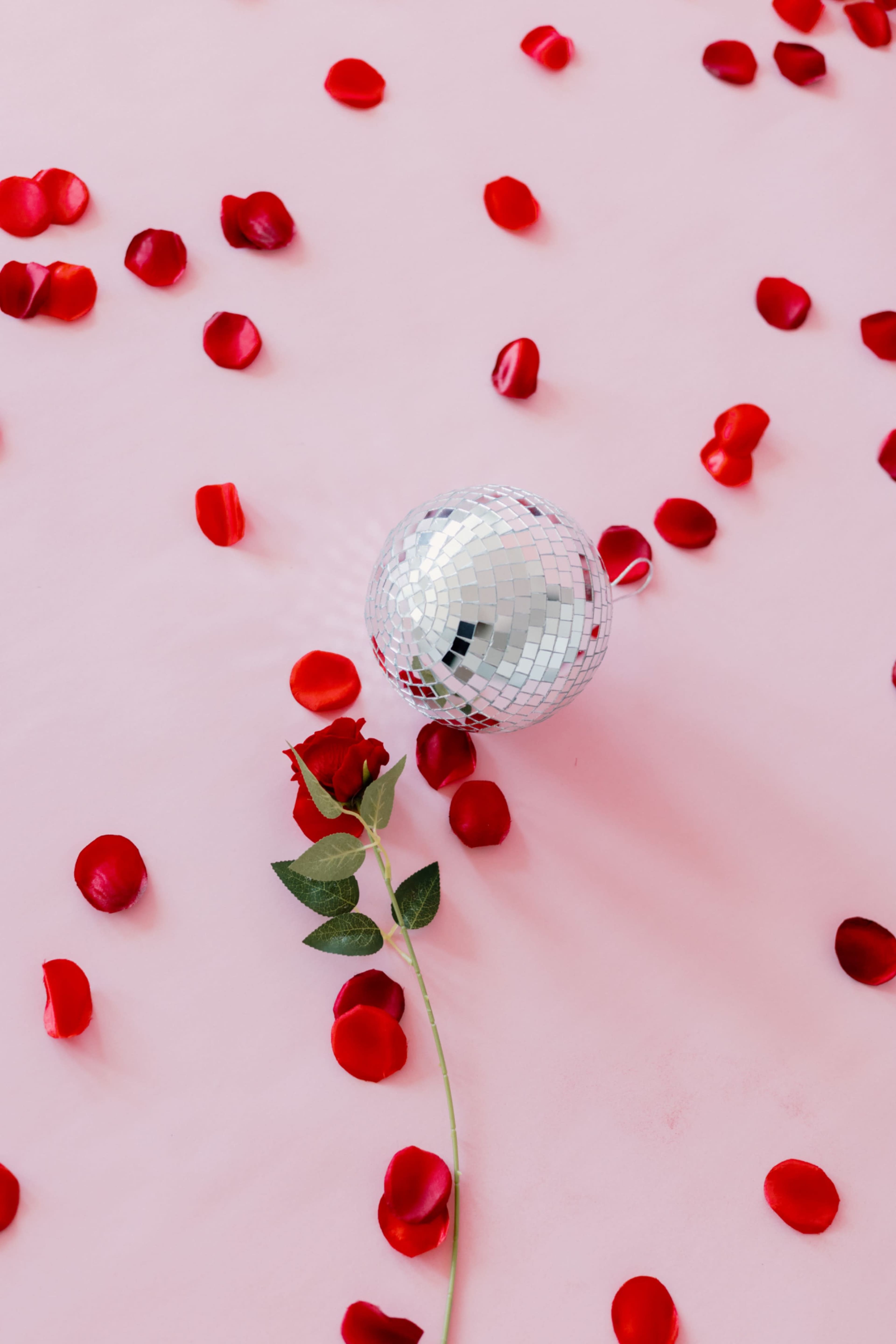 A silver disco ball rests on a pink surface surrounded by red rose petals and a single red rose with a green stem.