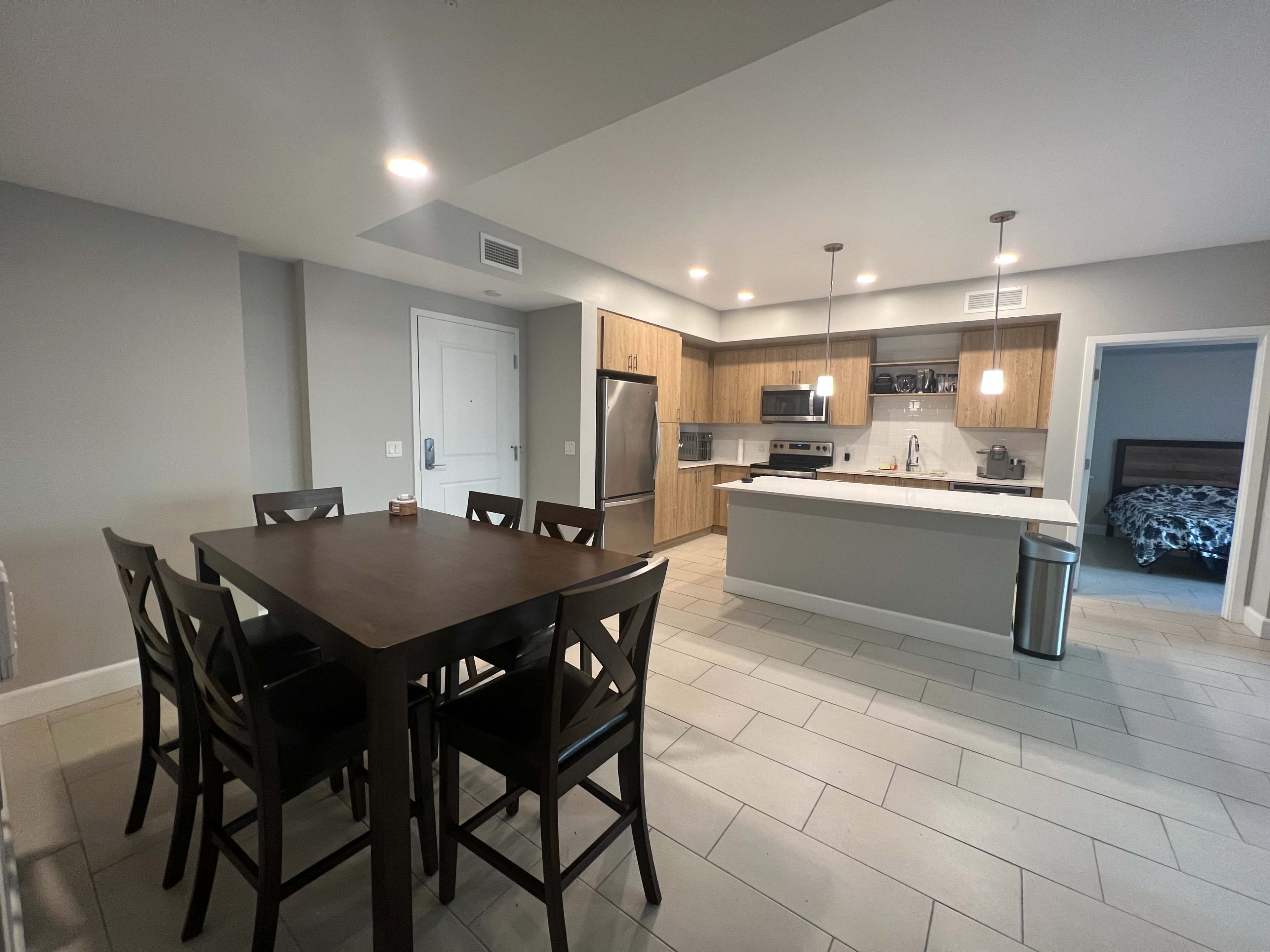 A modern kitchen and dining area with a wooden dining table, black chairs, and a kitchen featuring light wood cabinetry and stainless steel appliances.