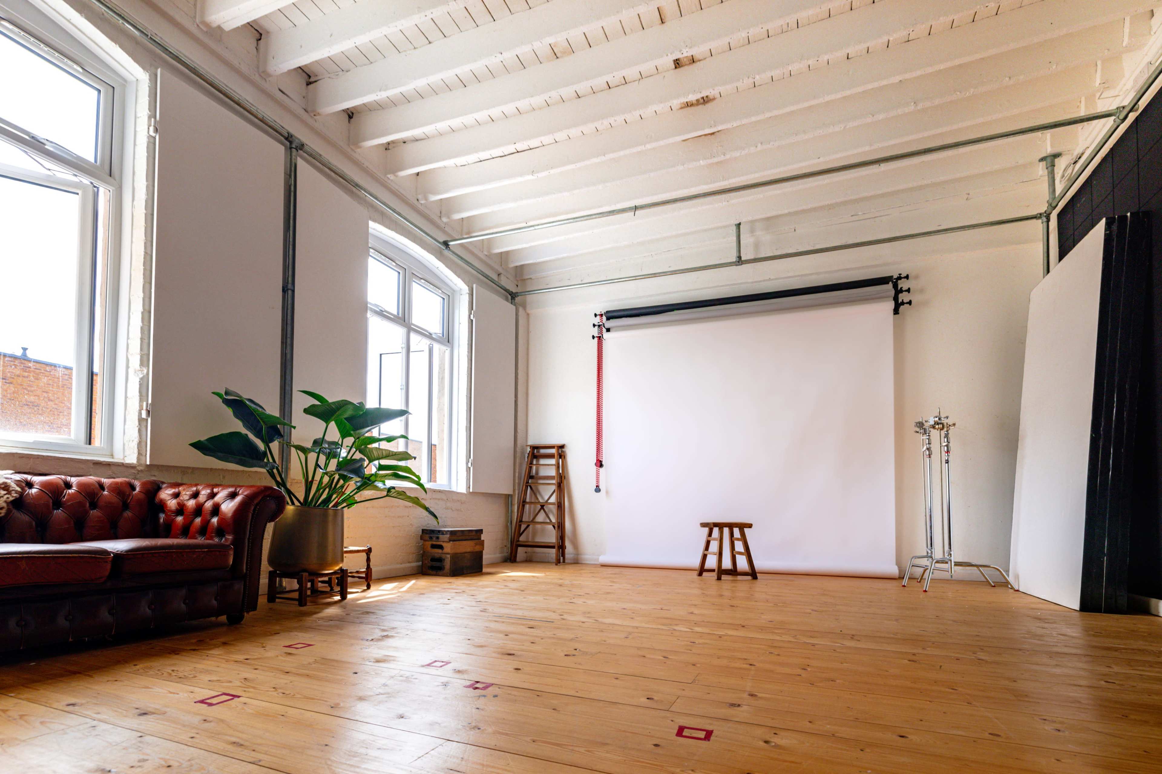 A spacious studio with high ceilings, a large white backdrop, a wooden floor, a brown leather couch, a stool, and a potted plant.