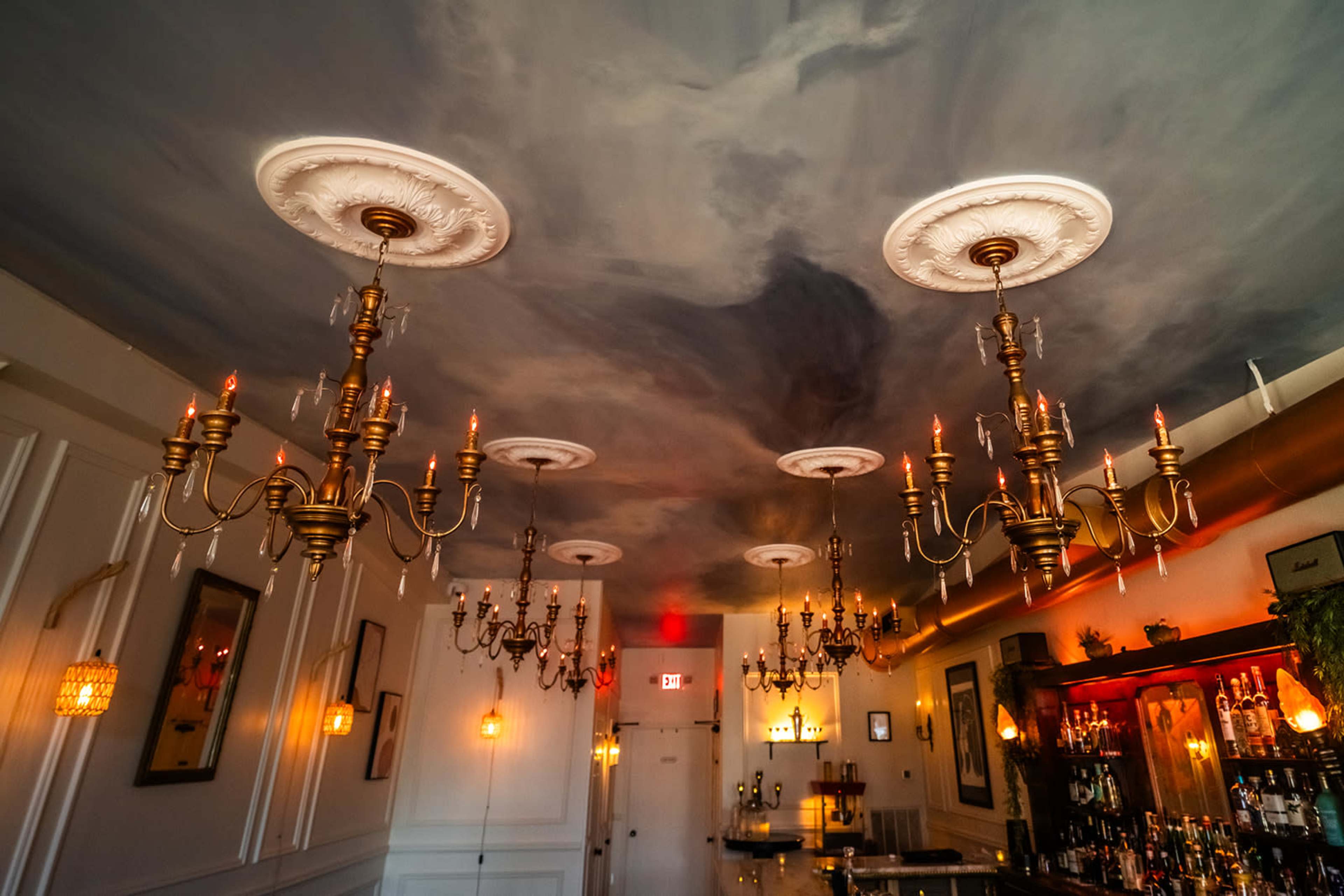 A dimly lit bar interior with ornate chandeliers hanging from a painted ceiling.