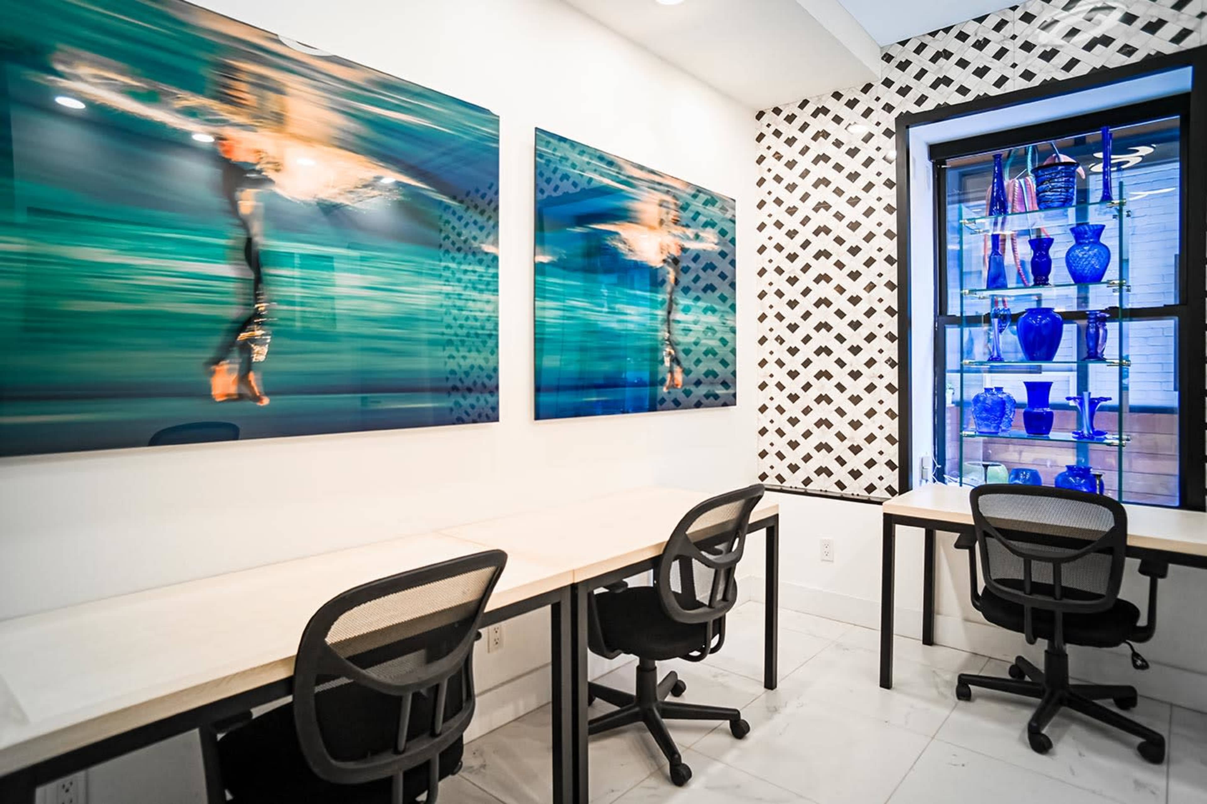 The image shows a modern office space featuring two large abstract artworks on the wall, two desks with chairs, and a display case filled with blue decorative vases.