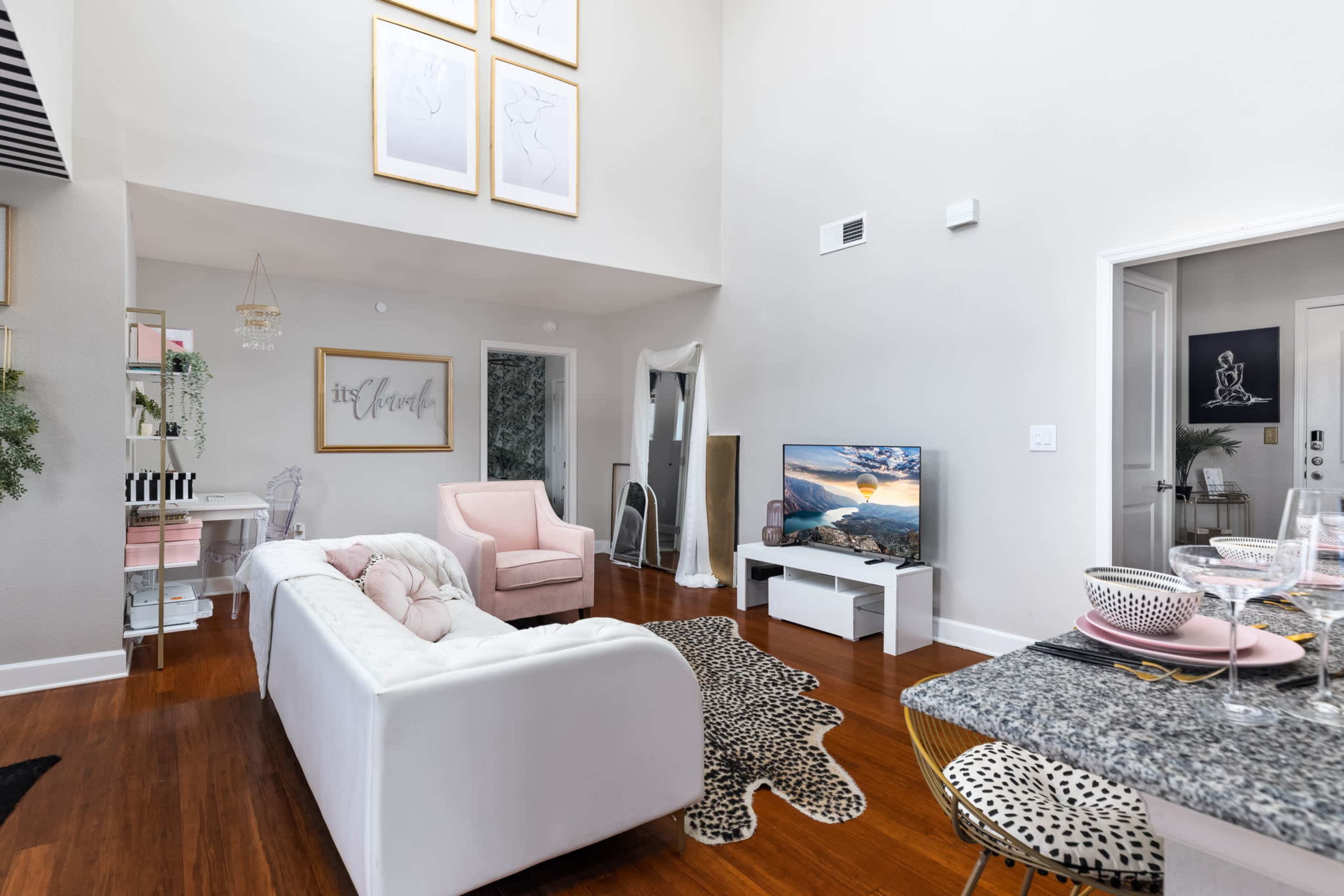The image shows a brightly lit living room featuring a white sofa, a pink accent chair, a large television, and a dining area with a granite countertop.