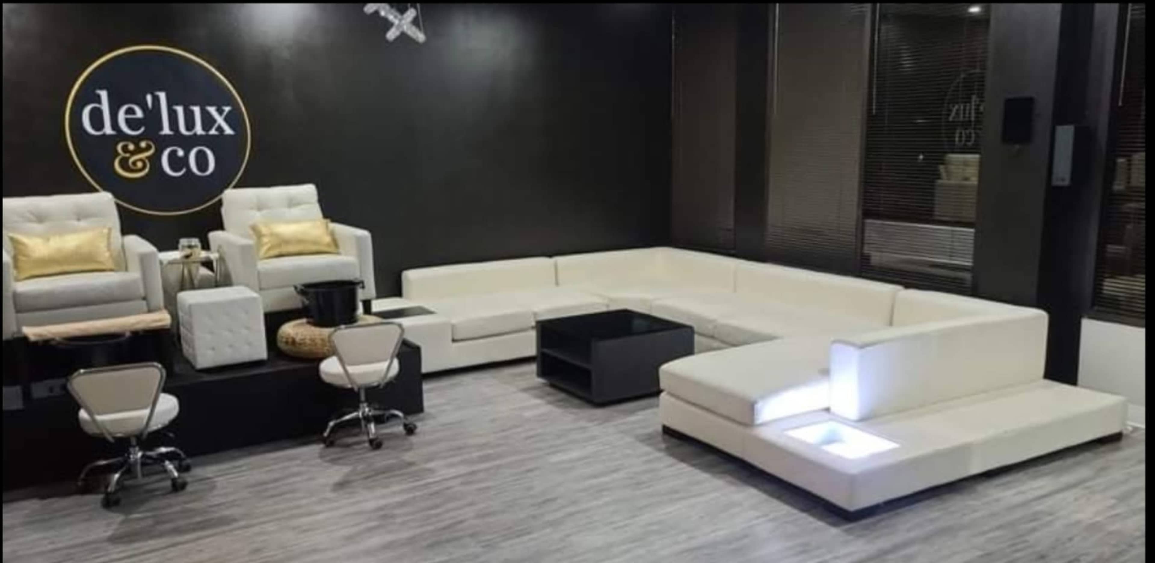A modern lounge area with white furniture, including a sectional sofa, two armchairs, and a black coffee table, set against a dark wall with a logo that reads "de/lux & co."