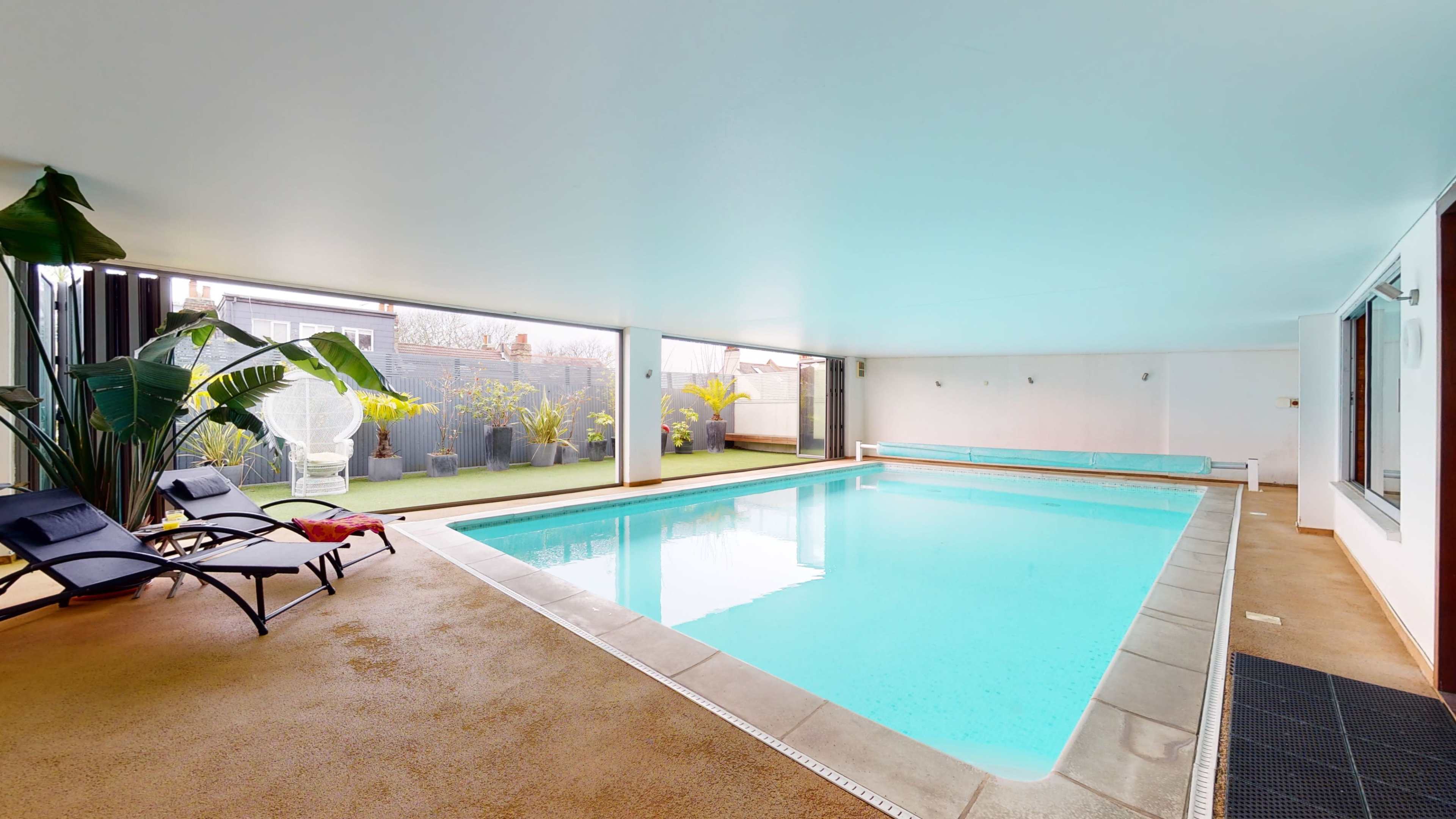 The image shows an indoor swimming pool area with lounge chairs, surrounded by large windows and potted plants.