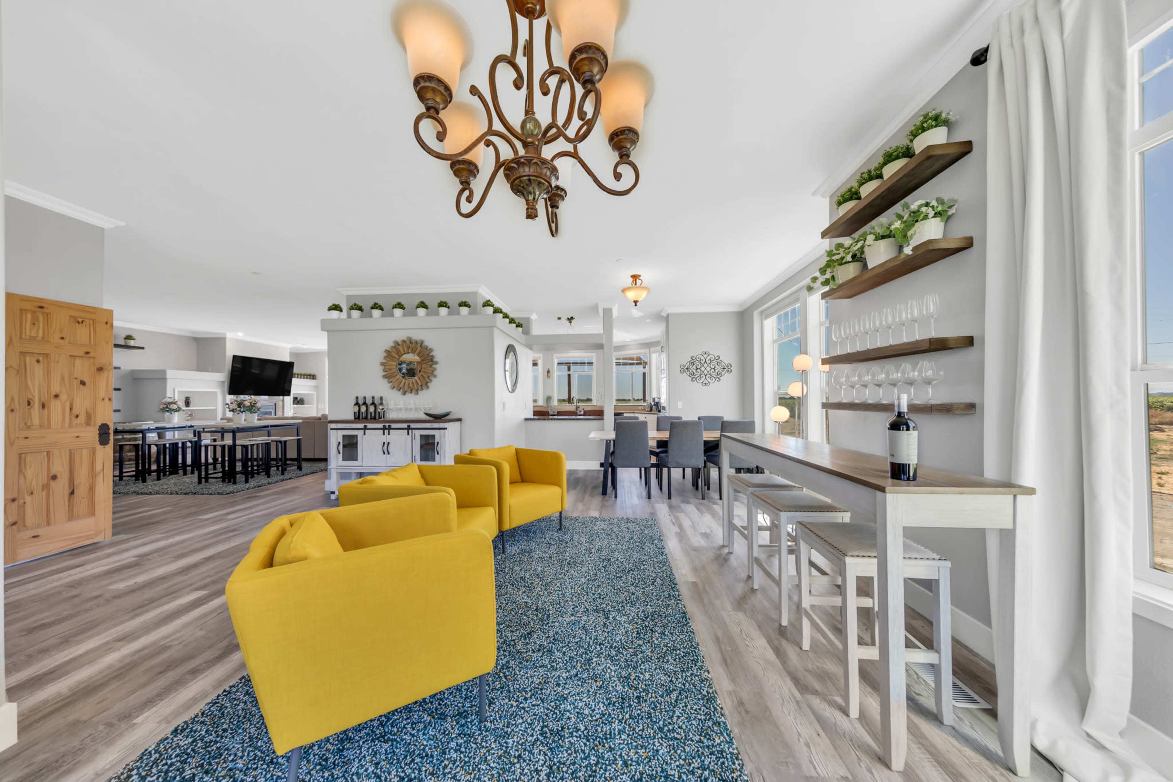 A brightly lit living area with two yellow armchairs, a patterned rug, a bar table with wine display, and an open view into a modern kitchen.