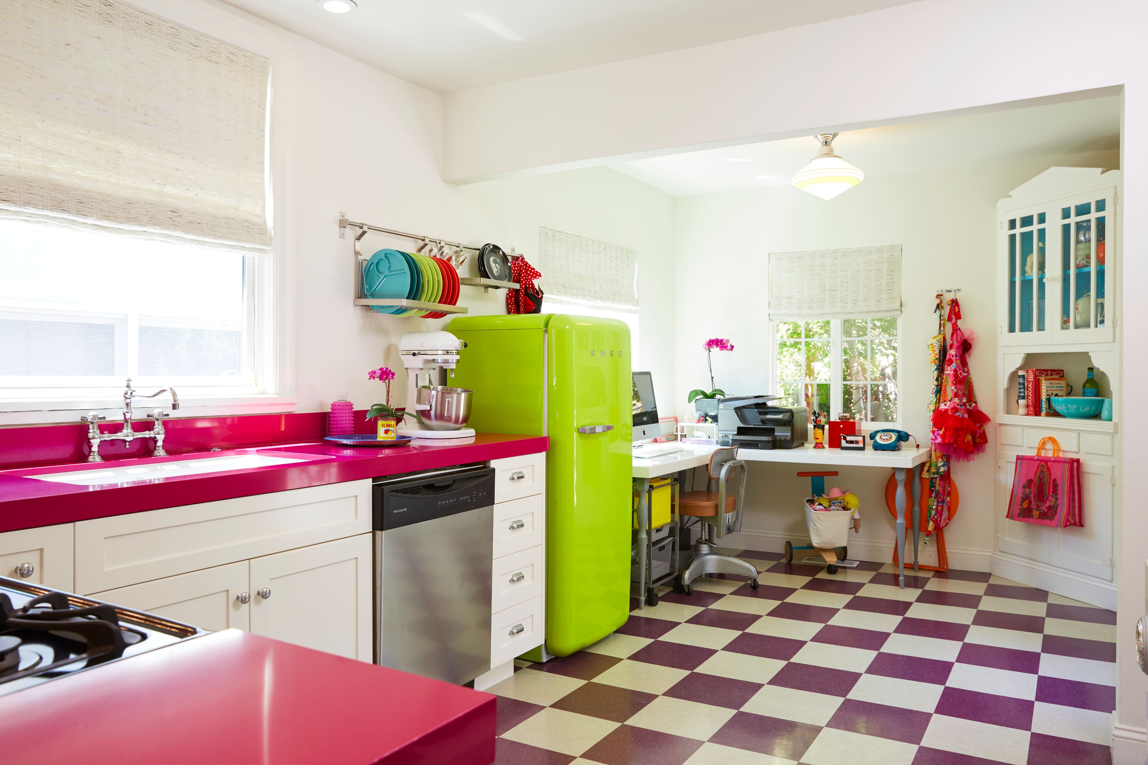 A brightly colored kitchen features a green refrigerator, pink countertops, and a checkered floor, with a workspace in the corner.