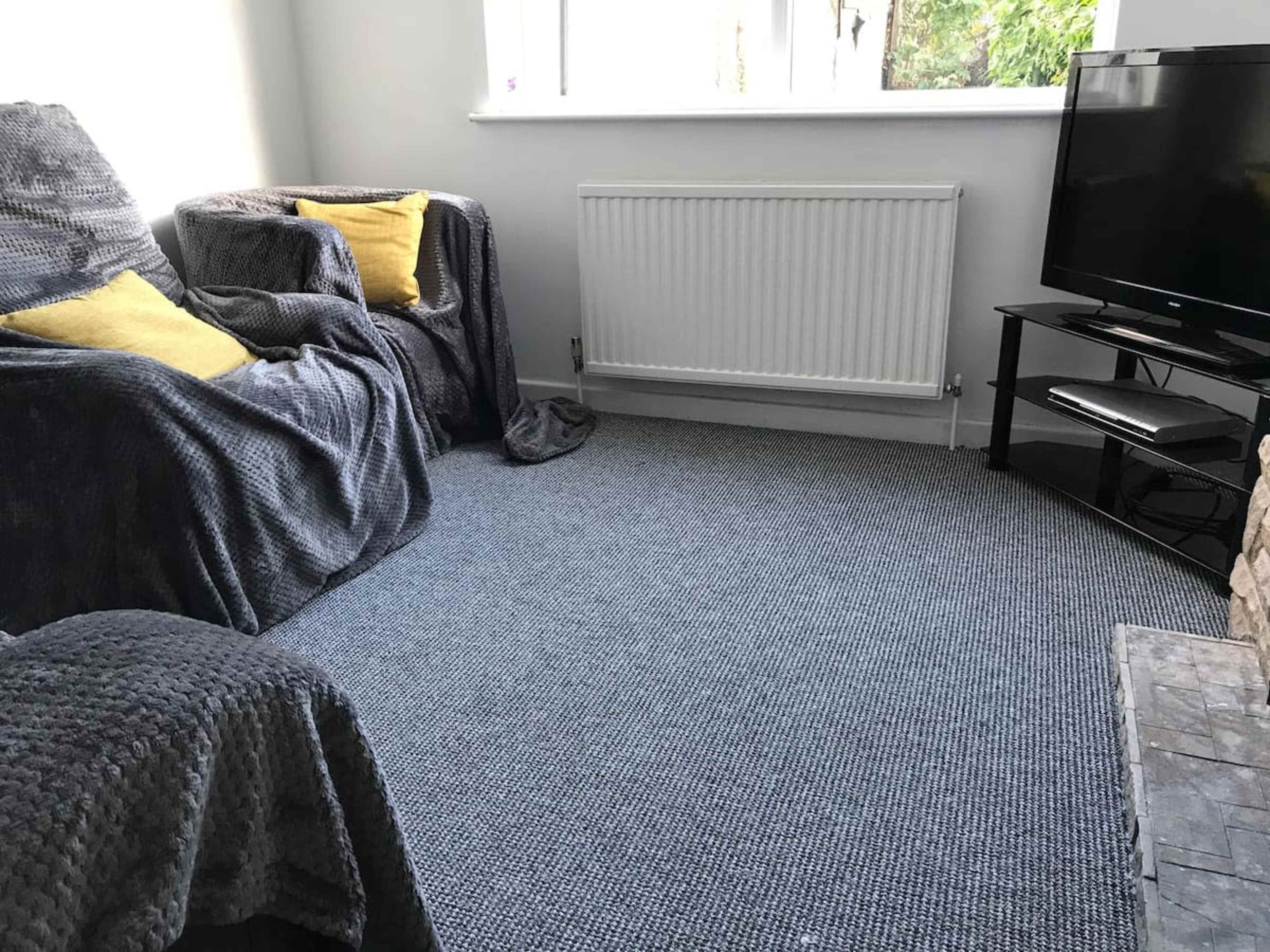 A cozy living room with a gray sofa covered in blankets, a yellow cushion, a radiator, and a black television stand.