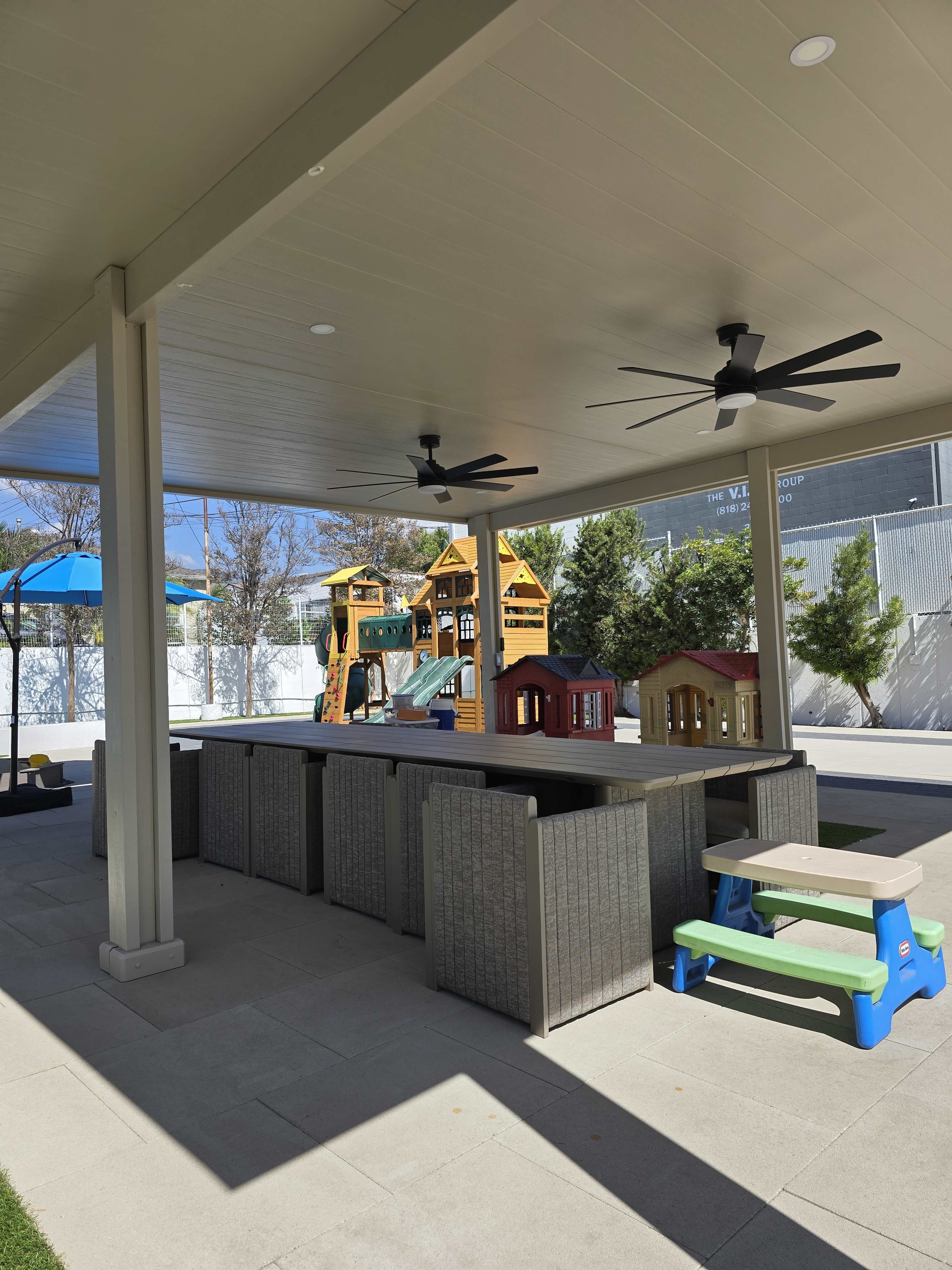 The image shows a covered outdoor space with picnic tables and playground equipment in the background.