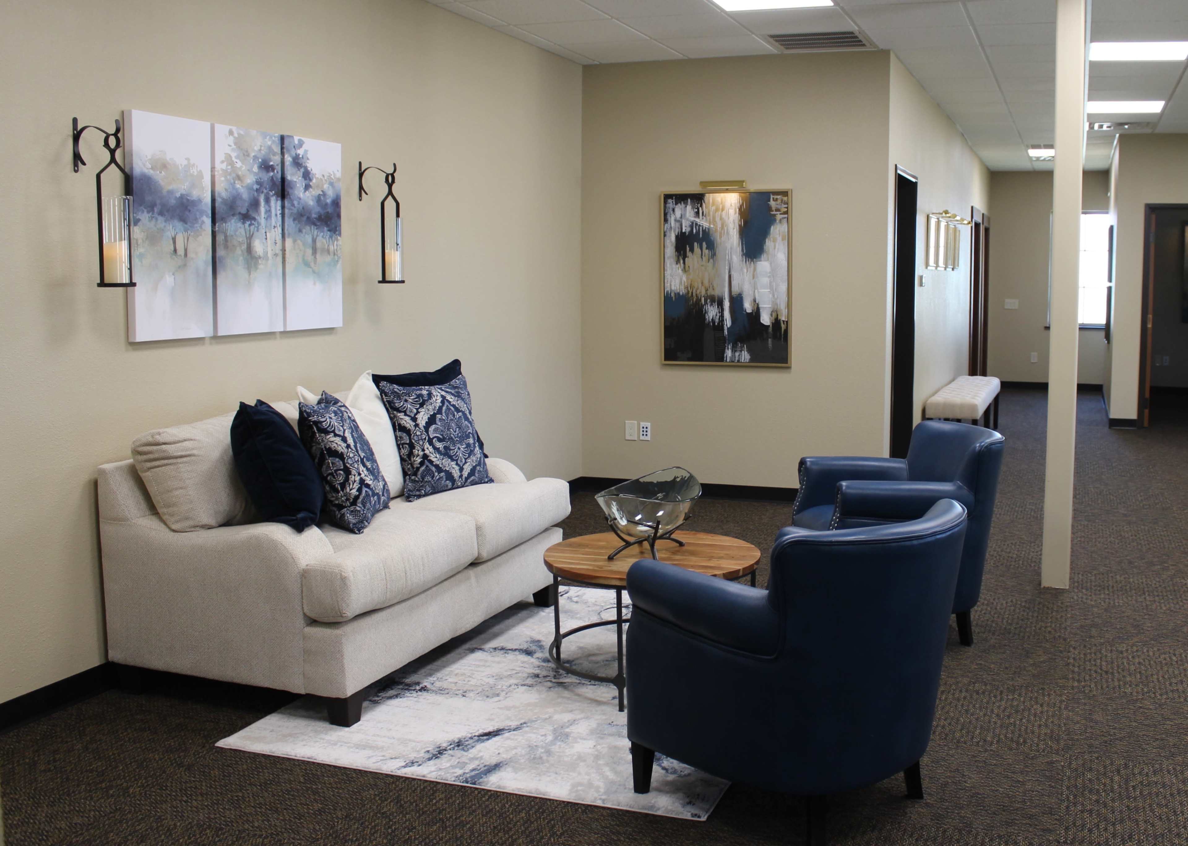 A modern office waiting area featuring a white sofa with blue and white patterned pillows, two blue armchairs, a wooden coffee table, and abstract wall art.
