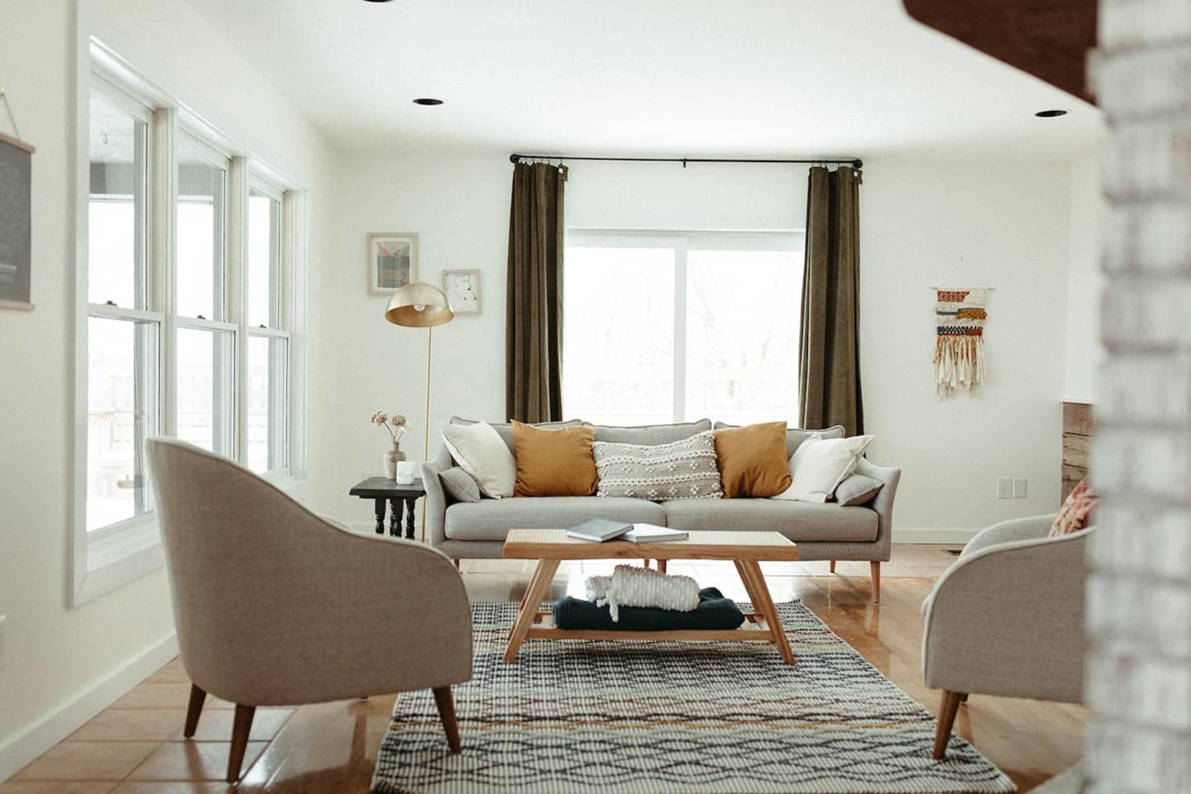 A modern living room features a gray sofa with decorative pillows, two armchairs, a coffee table, and large windows with curtains.