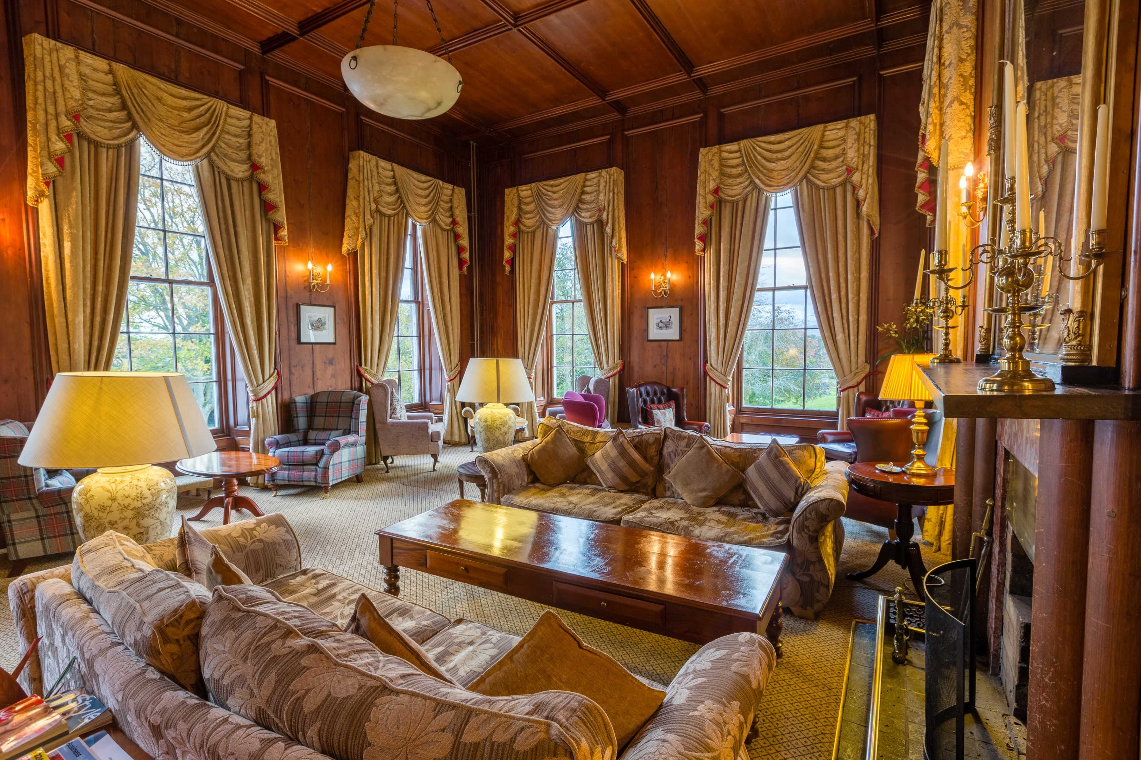 The image shows a cozy, well-furnished living room with large windows, wooden walls, and multiple seating areas, including sofas and armchairs.