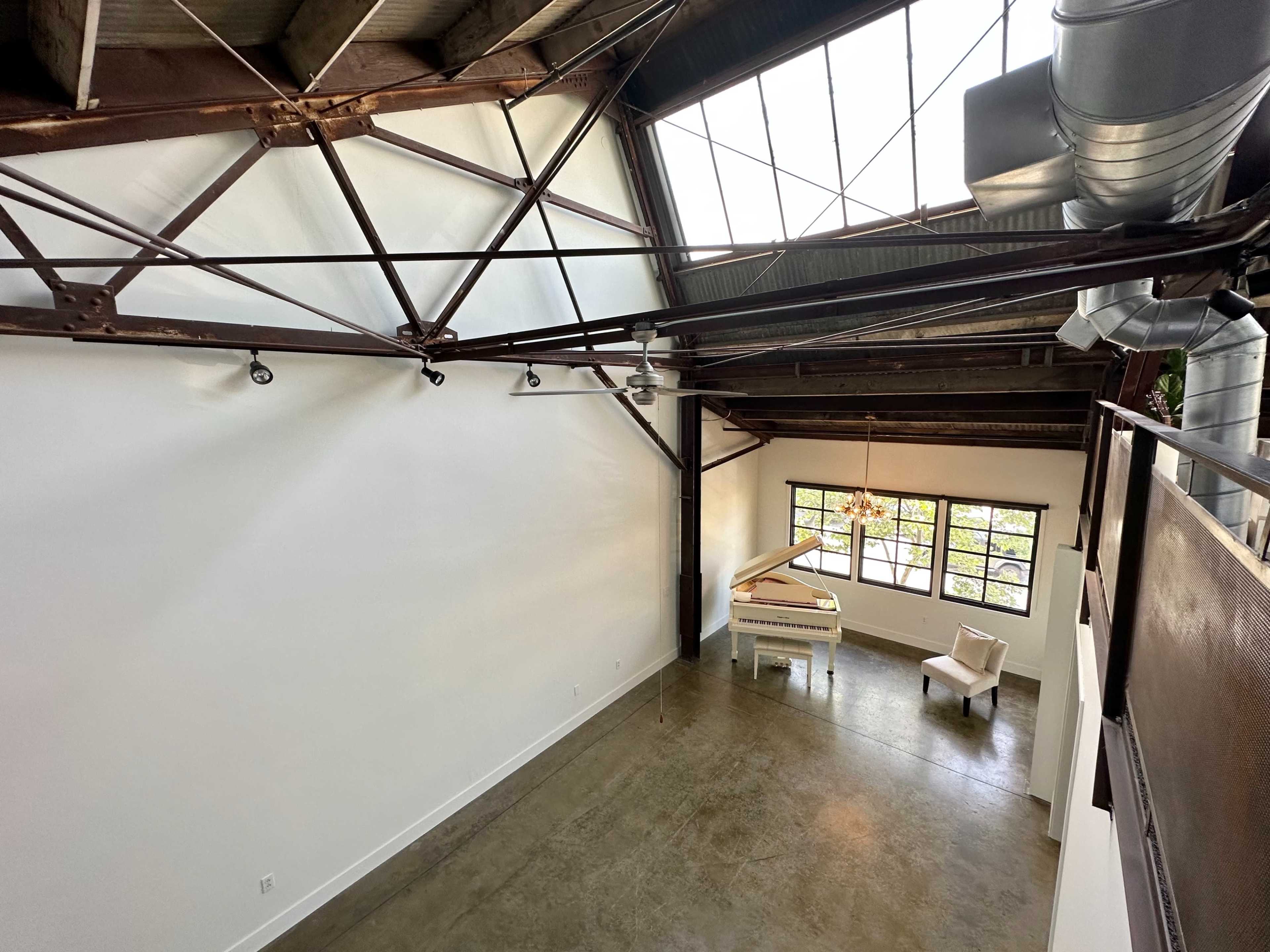 An open, industrial-style interior space with high ceilings, featuring a white grand piano, a chair, and large windows that allow natural light to enter.