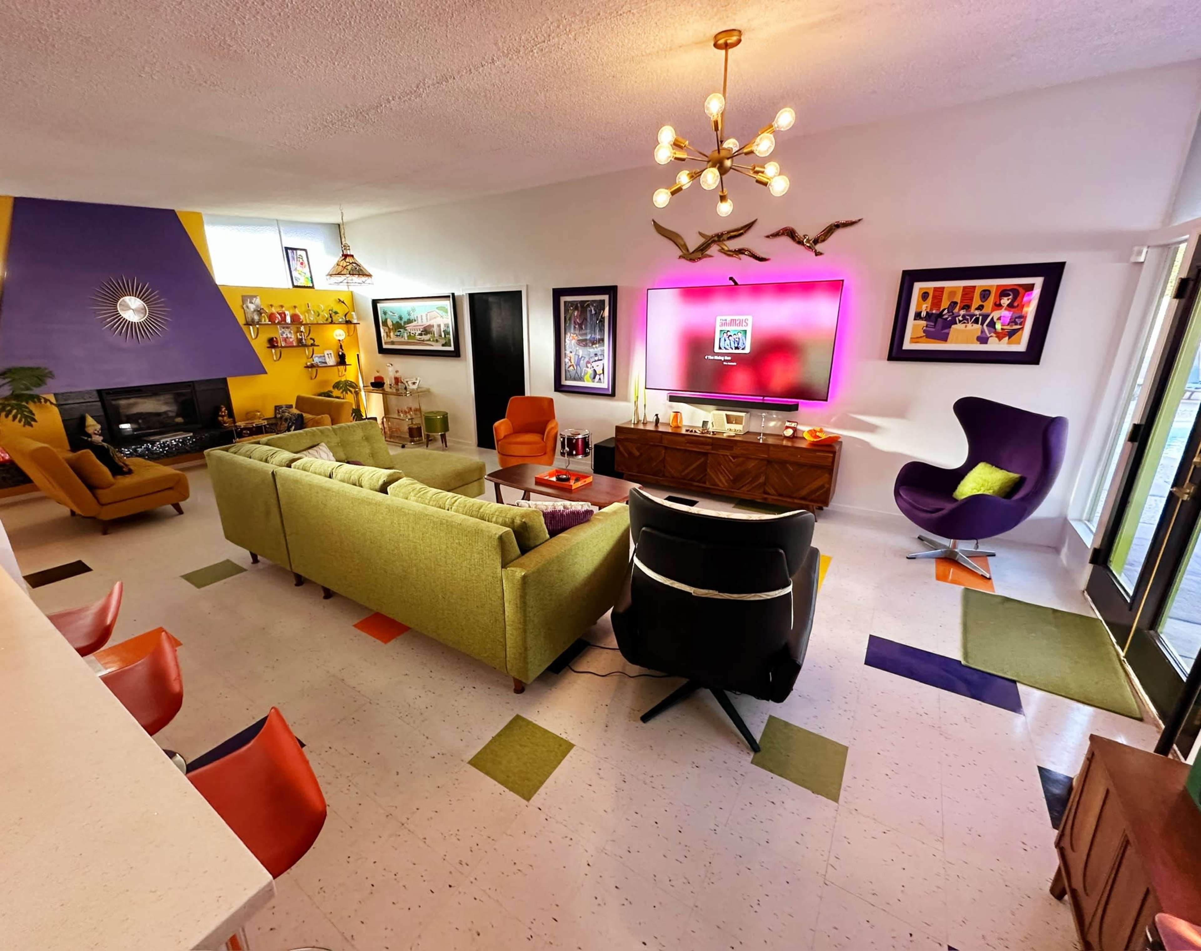 The living room features a variety of colorful furnishings, including a green sofa, purple accent chair, and a large wall-mounted television.