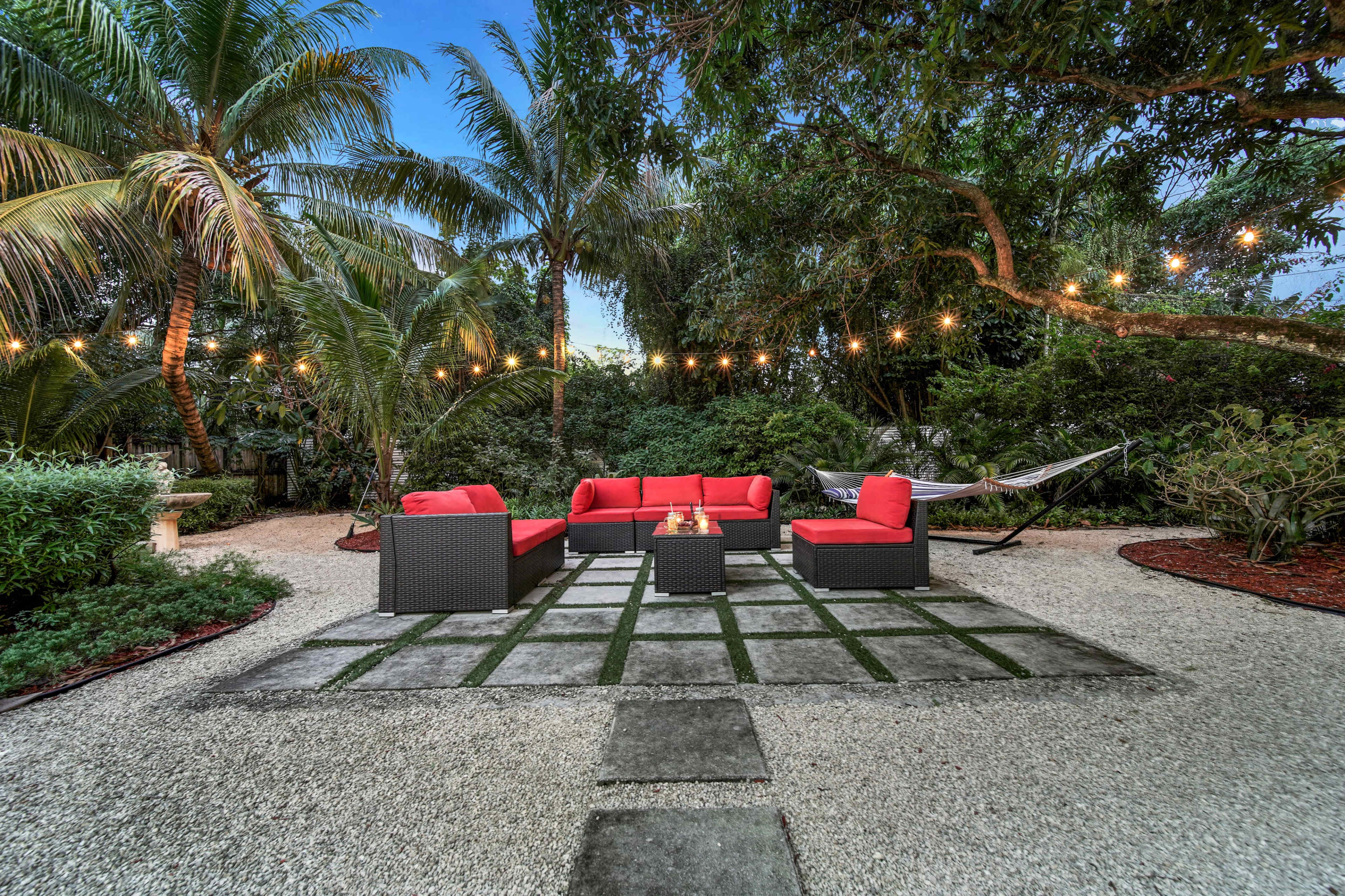 A landscaped outdoor space with a seating area of red cushions, a stone pathway, a hammock, and string lights among palm trees.