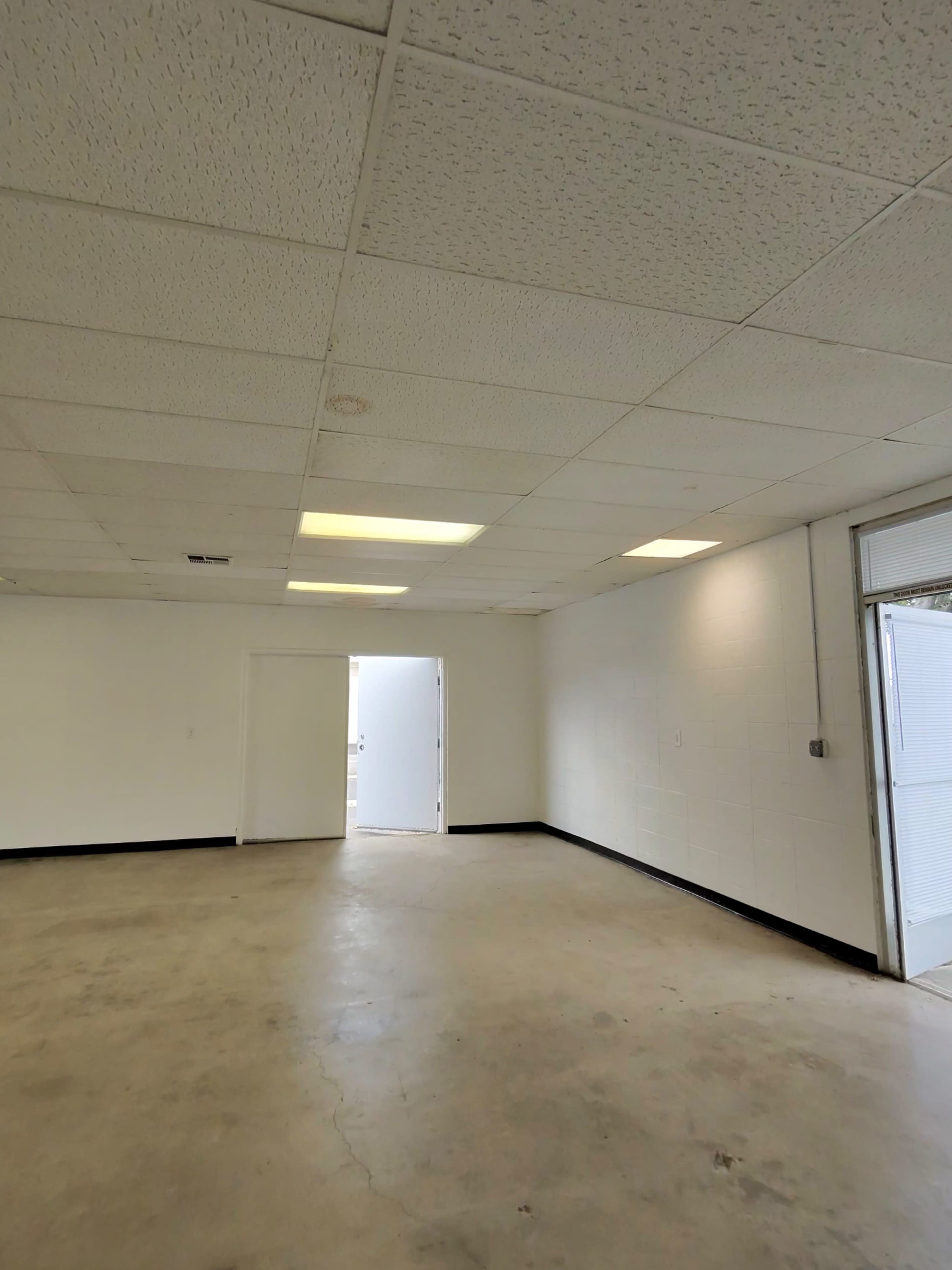 The image shows a spacious, empty room with a concrete floor, white walls, ceiling tiles, and a doorway leading to another area.