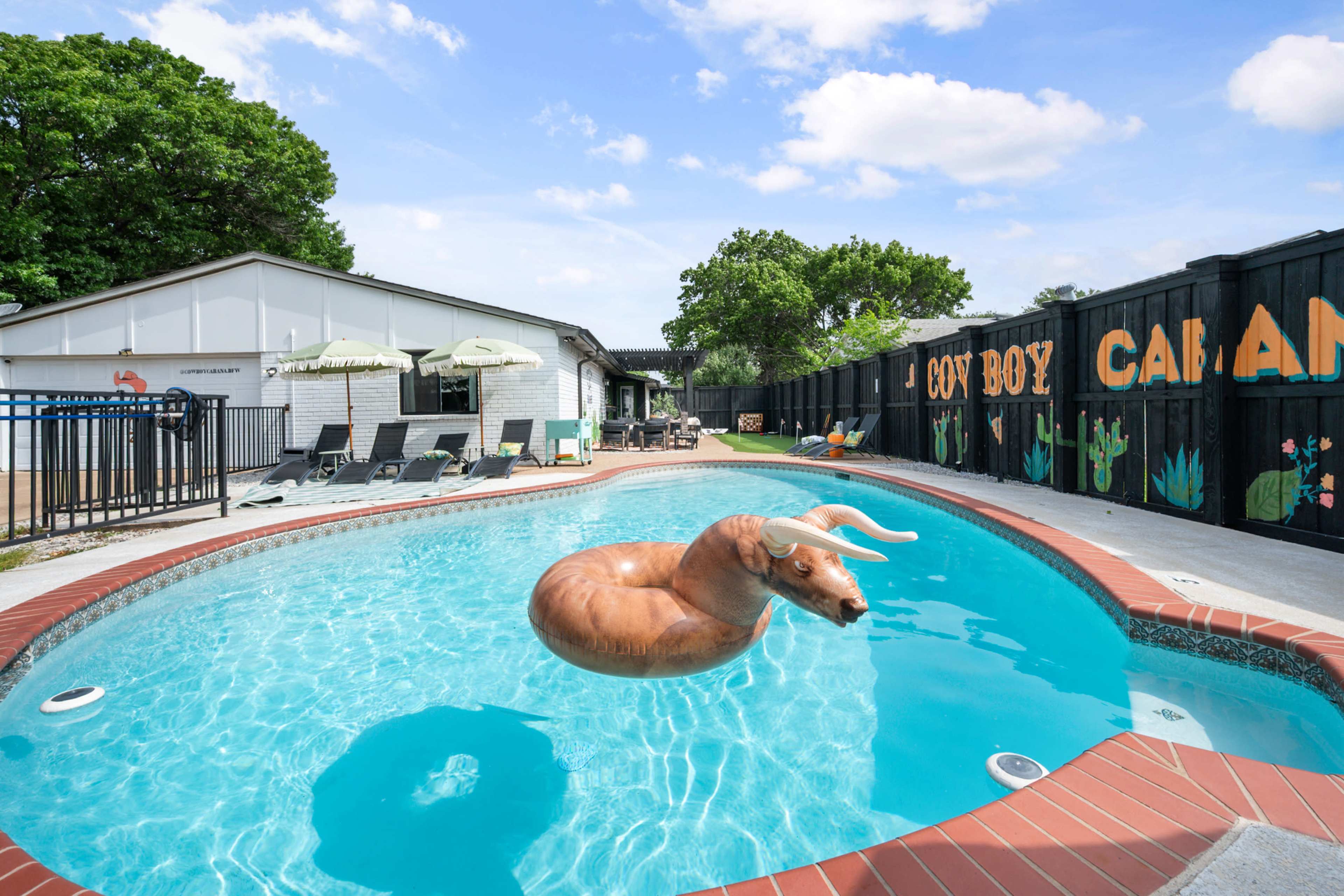 A pool with clear water features a large, inflatable goat float in the center, surrounded by lounge chairs and a building in the background.