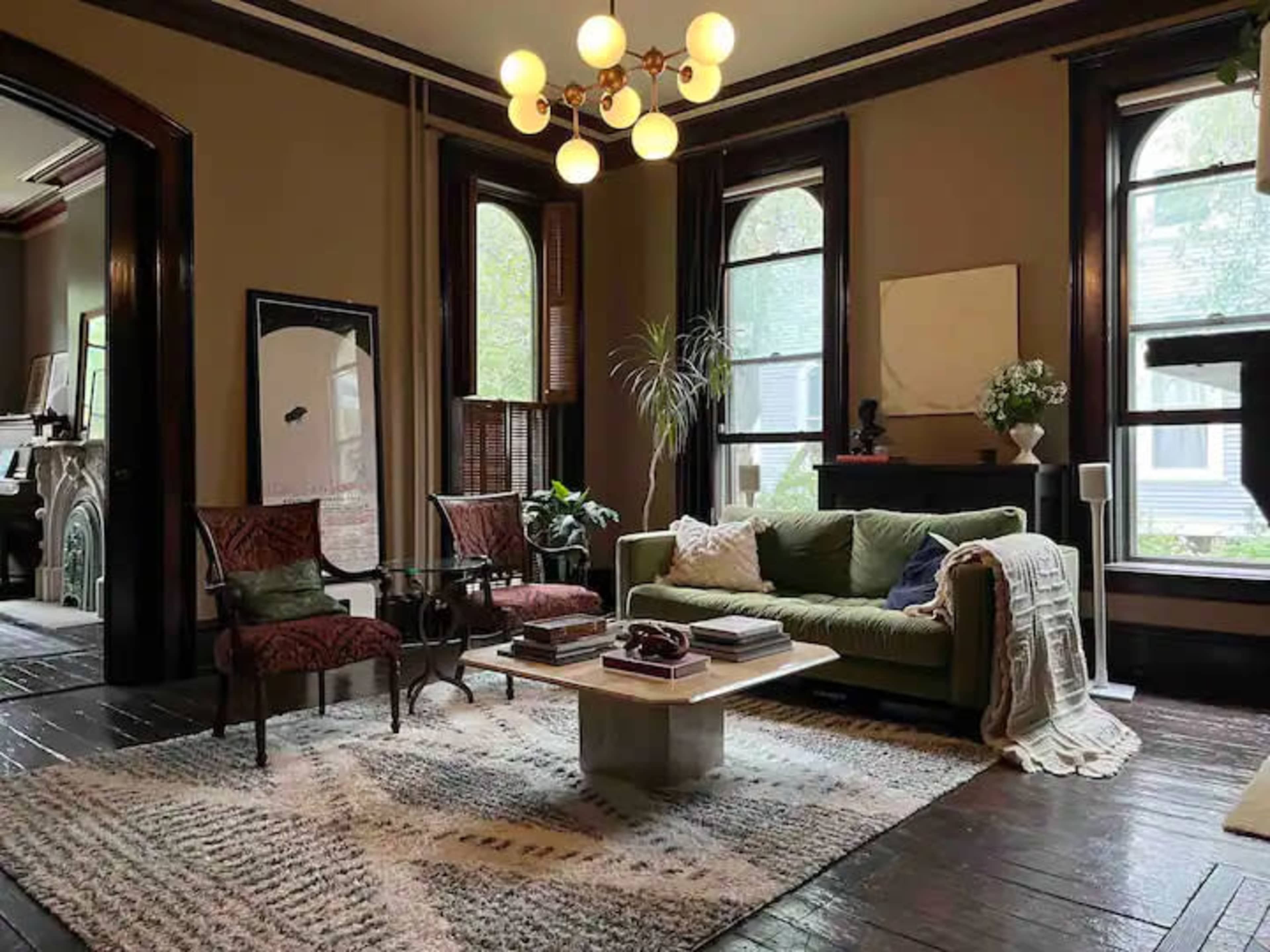 A well-furnished living room with a green sofa, a glass coffee table, and two chairs, illuminated by a chandelier, featuring large windows and a patterned rug.