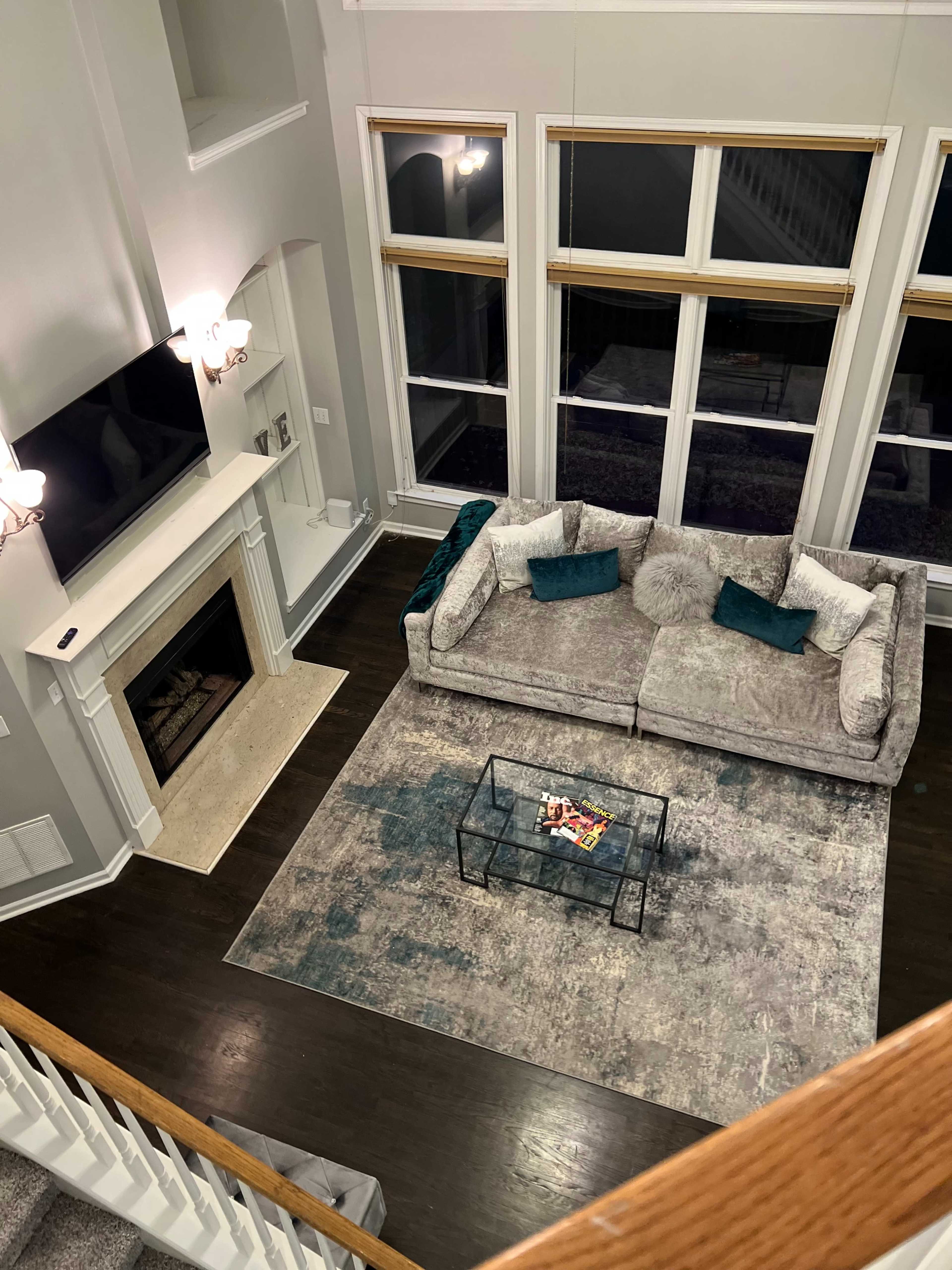 A spacious living room features a large gray couch, a glass coffee table, and a modern fireplace beneath a wall-mounted television, surrounded by tall windows.