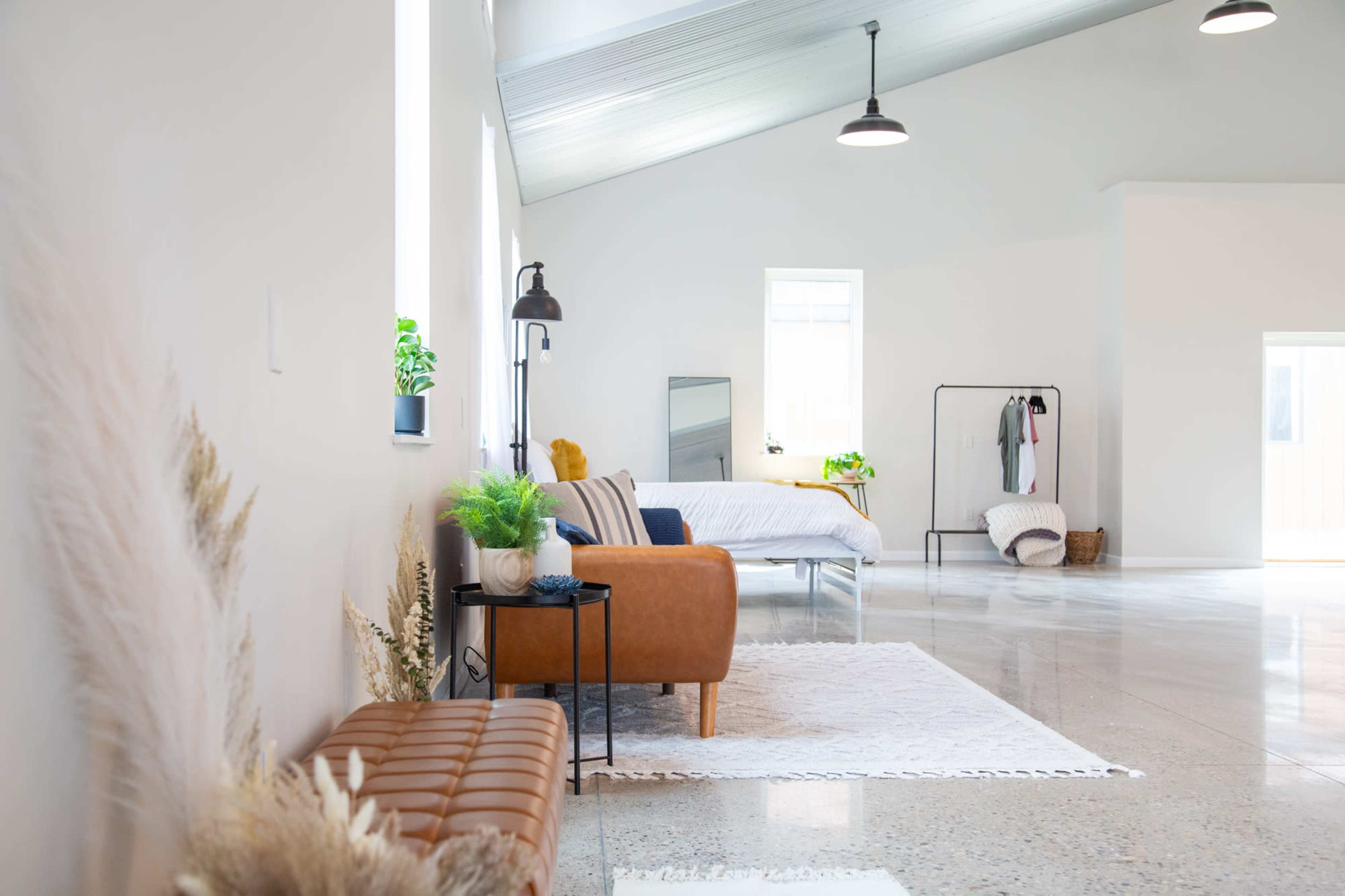 A spacious, minimalistic bedroom features a bed, a leather sofa, decorative plants, and a clothing rack in a well-lit room with concrete flooring.