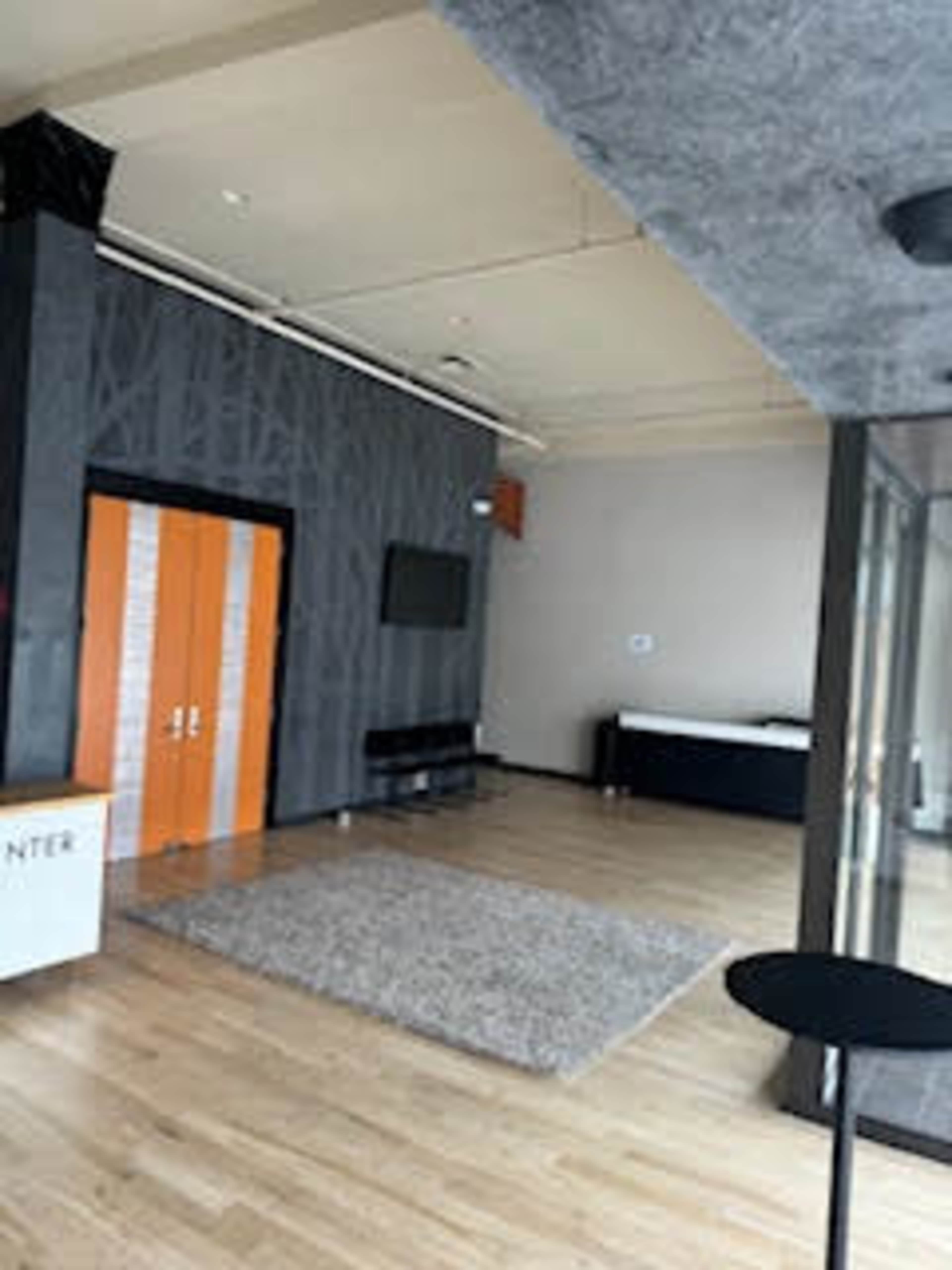 The image shows an empty, modern interior space with wooden flooring, a large area rug, and a seating area, featuring orange double doors and a television mounted on the wall.