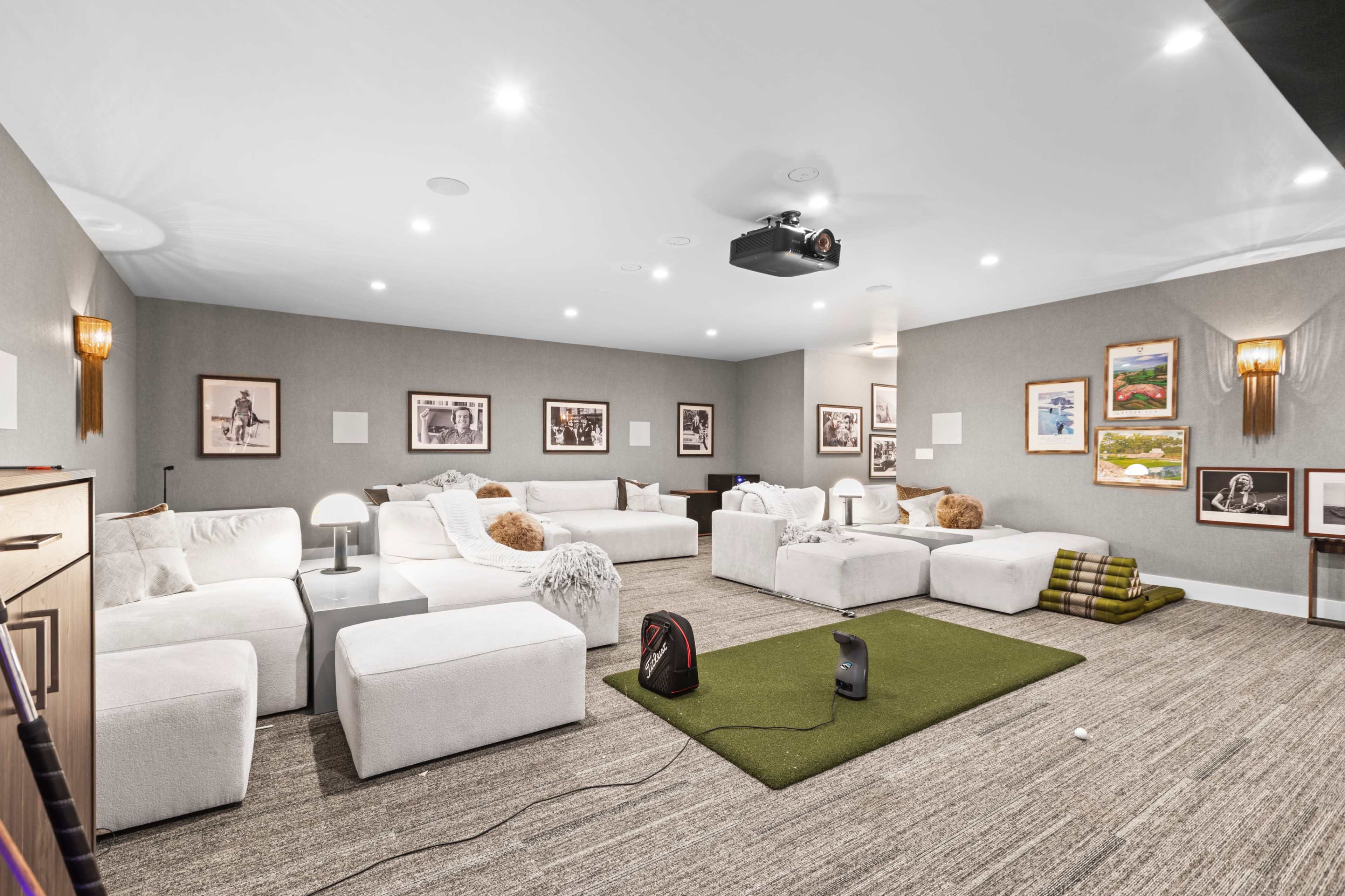 A spacious media room with white sofas, a projector on the ceiling, and artwork displayed on the walls.