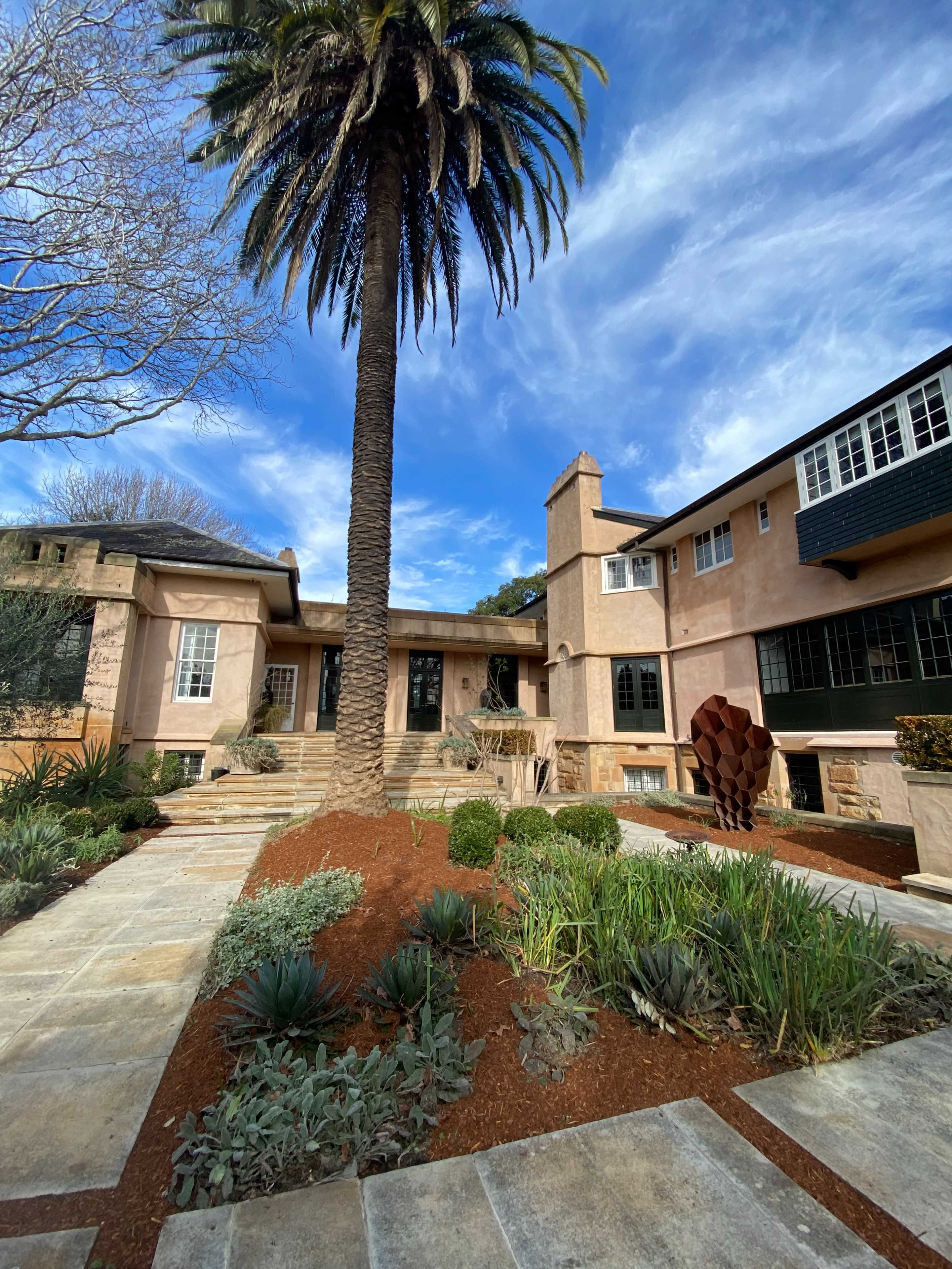 A large, pink stucco house with a palm tree and a landscaped garden featuring stone pathways and a sculpture.
