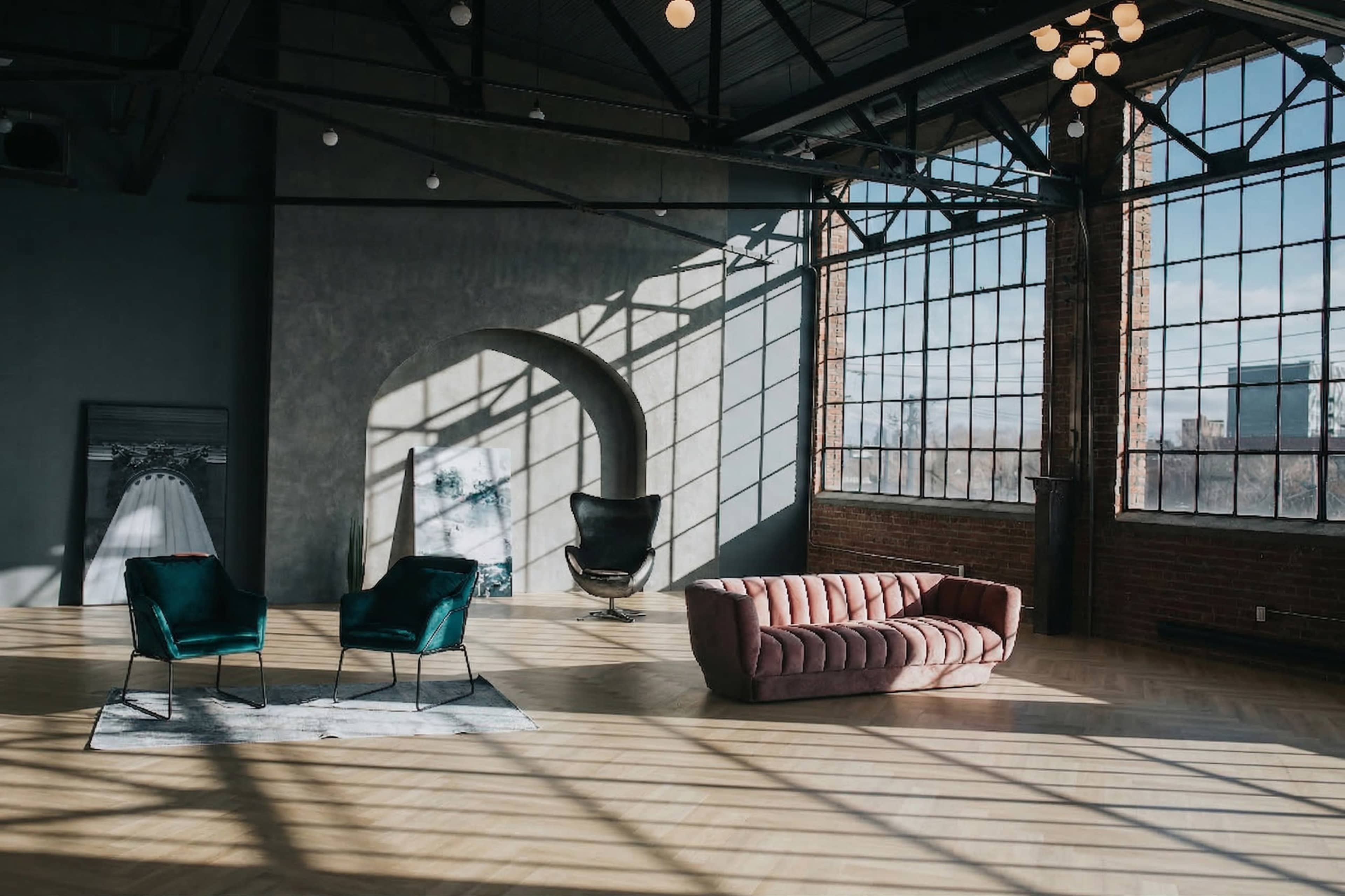 The scene features a spacious, modern loft with large windows, showcasing two green armchairs, a pink sofa, and natural light casting shadows on the wooden floor.