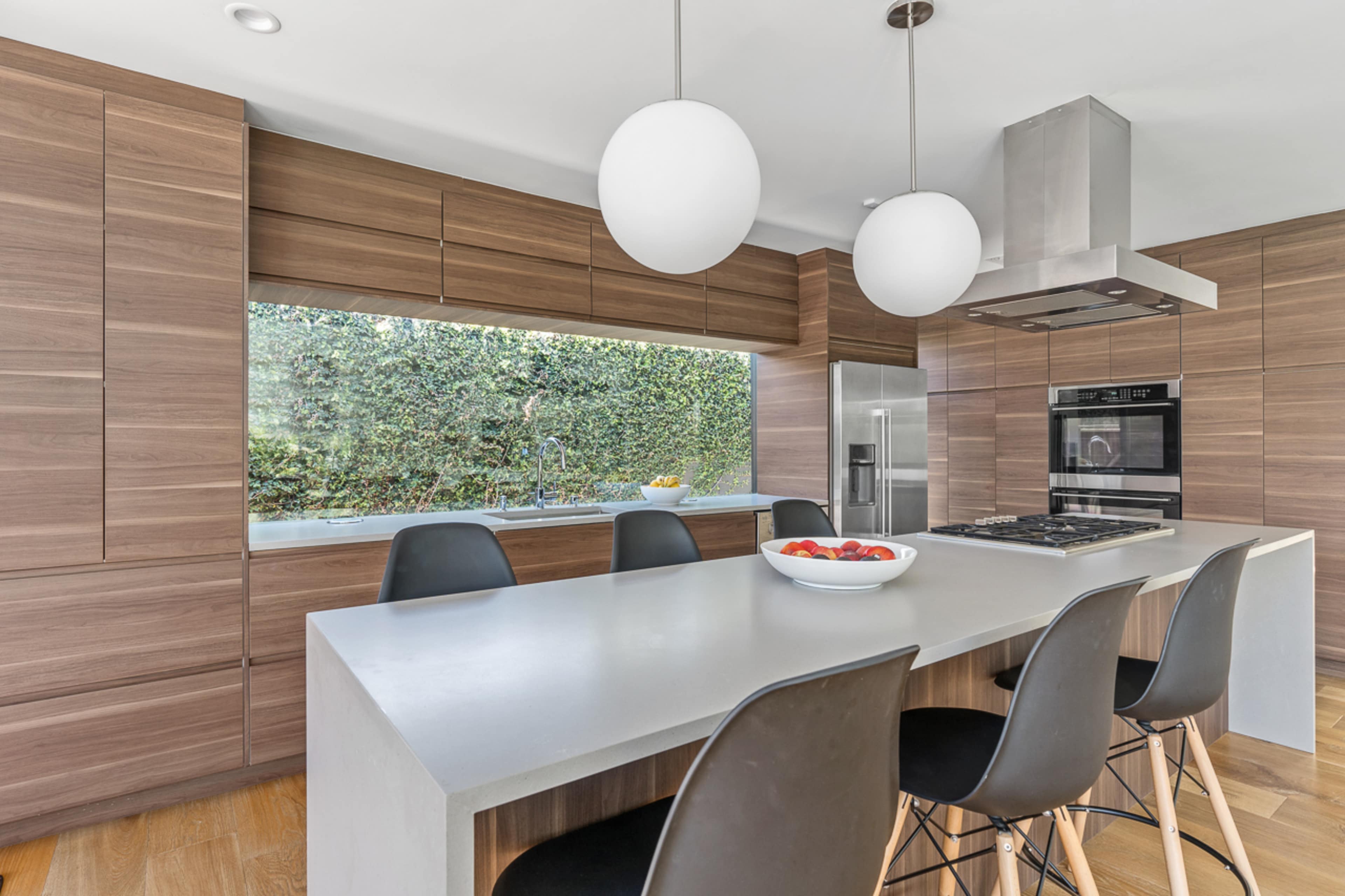 The kitchen features sleek wooden cabinetry, a large island with seating, and a window showcasing a view of greenery outside.
