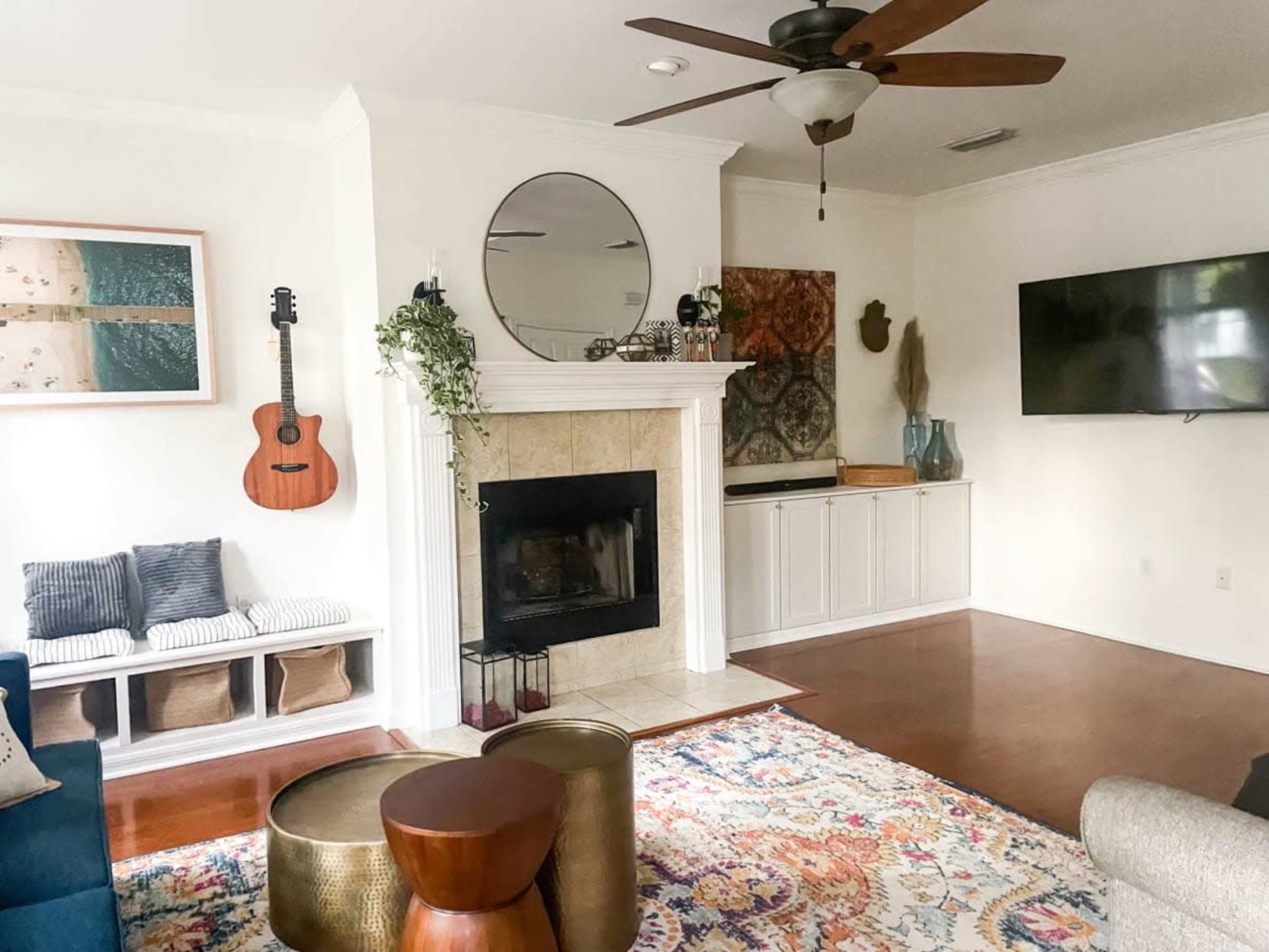 The living room features a fireplace with a circular mirror above it, accented by a guitar and a large television mounted on the wall, along with a colorful area rug and various decorative elements.
